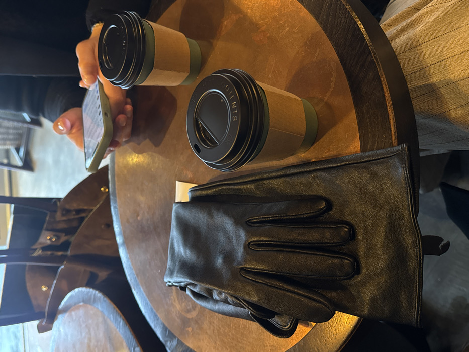 A round table holds two disposable coffee cups with black lids and cardboard sleeves. A pair of black leather gloves rests beside one of the cups. A person’s hand holding a phone is visible near the other cup. The table has a dark trim and a metallic sheen. The background includes clothing and a chair.