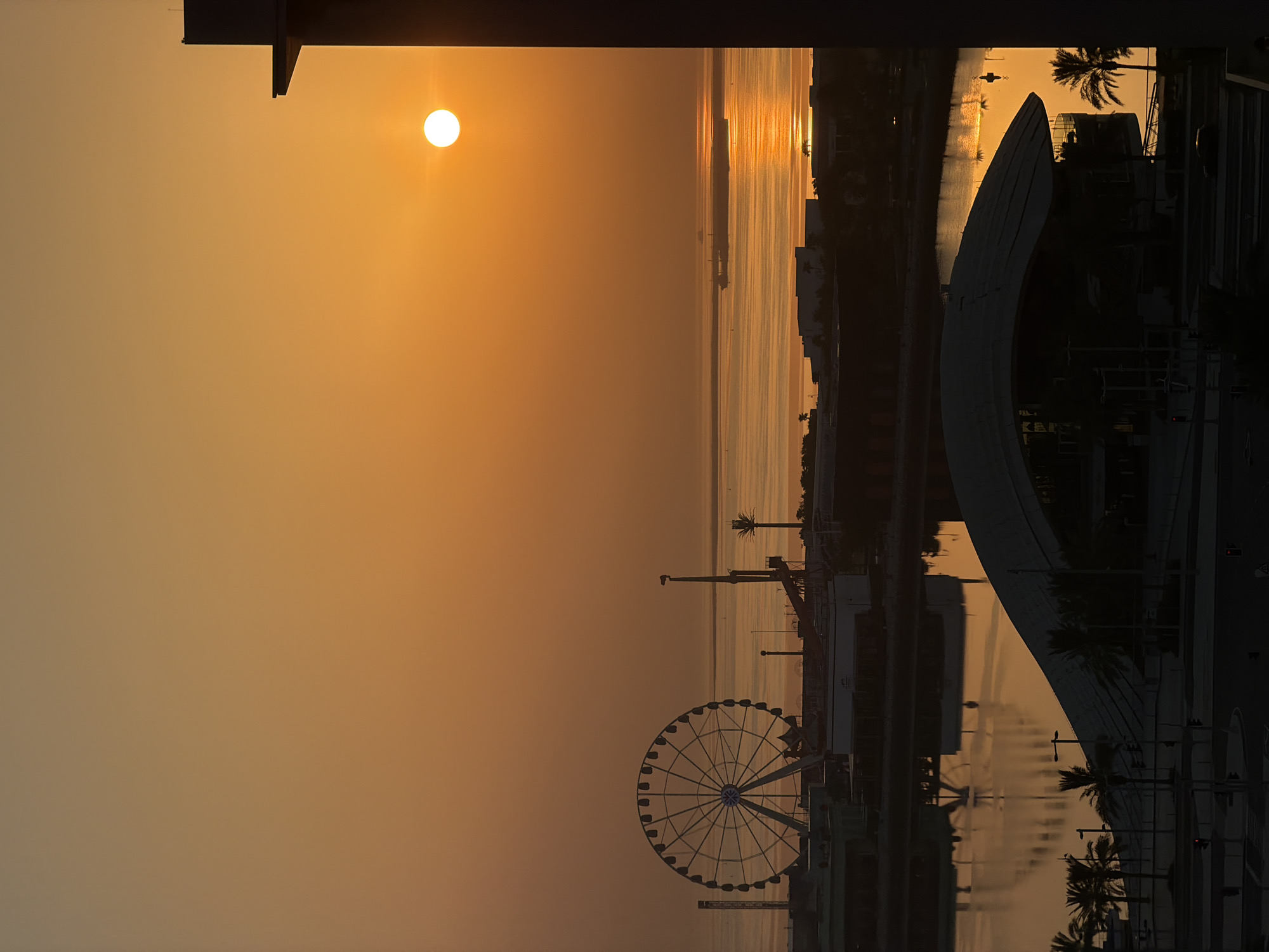A wide shot captures a silhouette of the Lusail waterfront at sunset. The sun, a bright orange orb, is positioned in the upper right quadrant, casting a warm glow across the sky and reflecting on the calm waters of the Arabian Gulf. In the distance, a large cargo ship is visible on the horizon. Closer to shore, a Ferris wheel stands prominently on the left side of the frame, a common fixture in waterfront amusement areas. Other amusement park structures and buildings are silhouetted against the sky. In the foreground, the distinctive curved roof of a modern building is visible, along with palm trees and streetlights. The overall scene depicts a peaceful evening at a city's recreational hub.