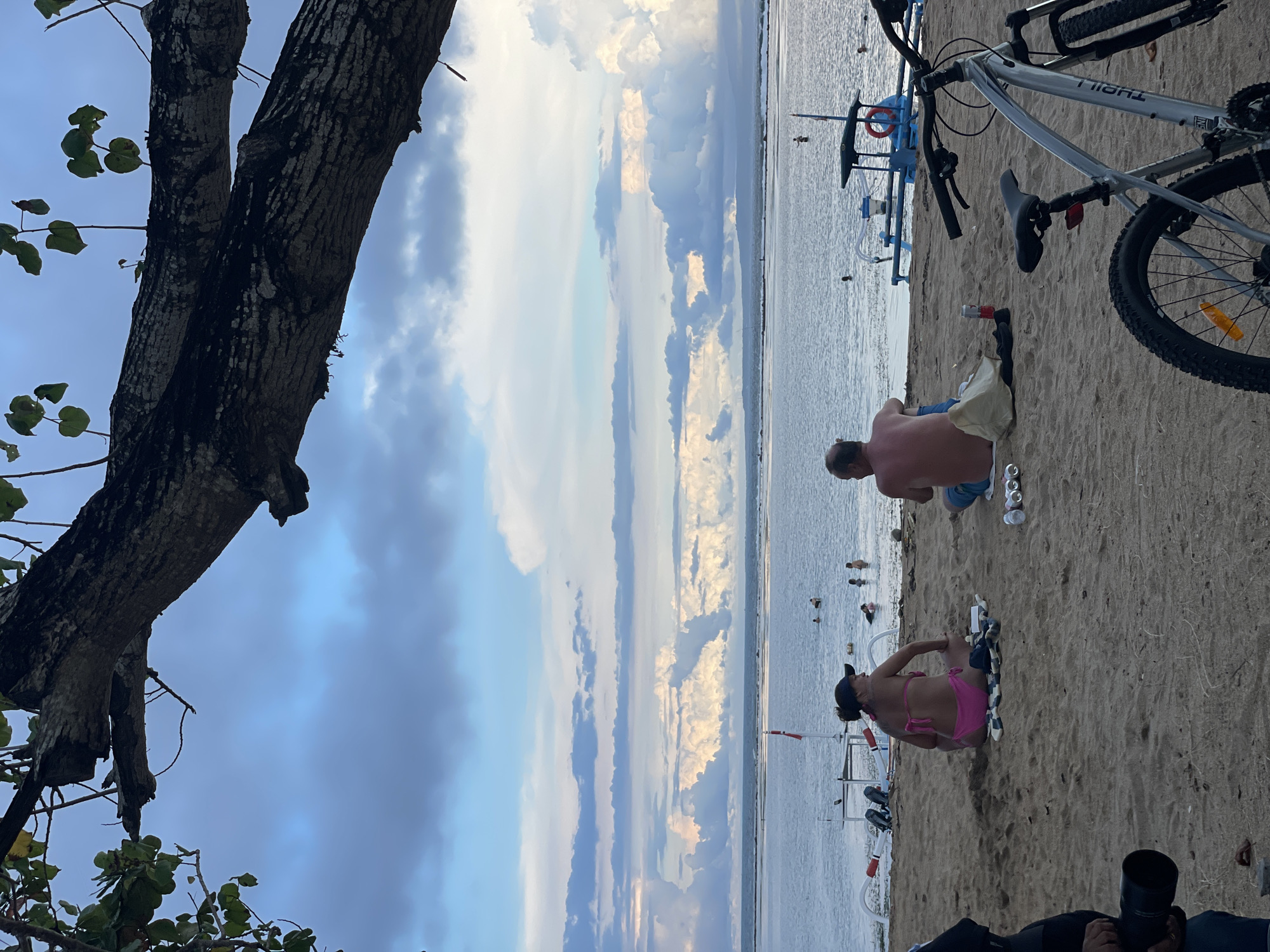 On a sandy beach in Denpasar, Indonesia, a scene unfolds with several focal points. In the lower right corner, a person in dark clothing holds a large camera with a lens, pointed towards a woman who is positioned prone on the beach in a pink bikini. Nearby, a man in blue shorts is seen sitting and looking towards the ocean. To their right, a bicycle with a silver frame stands upright on the sand. In the background, people are seen wading and swimming in the ocean near numerous boats. In the upper left corner of the photo, a tree branch with a textured bark extends into the scene. Overhead, the sky displays a mix of white and grey clouds. The shoreline stretches into the distance, creating a dynamic and layered composition.