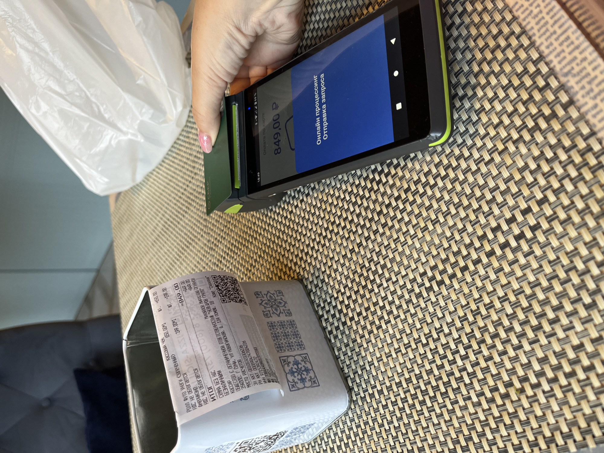 A hand with manicured nails inserts a green Sberbank card into a black and green payment terminal, which displays "849,00 Р" and "Online processing. Sending request." The terminal is positioned on a woven, beige and brown placemat. To the left of the terminal, a receipt from "KAFFEE "SOCHI"" is partially visible, extending from a metal container. The receipt lists items such as "ROLL FUTO VUHGZ (ZAZHELENNOY)" and "AMERIKANO," with corresponding prices. The background shows a blurred interior with a dark-colored sofa. The scene appears to be in Sochi, Russia, suggested by the text on the receipt and the Sberbank card.