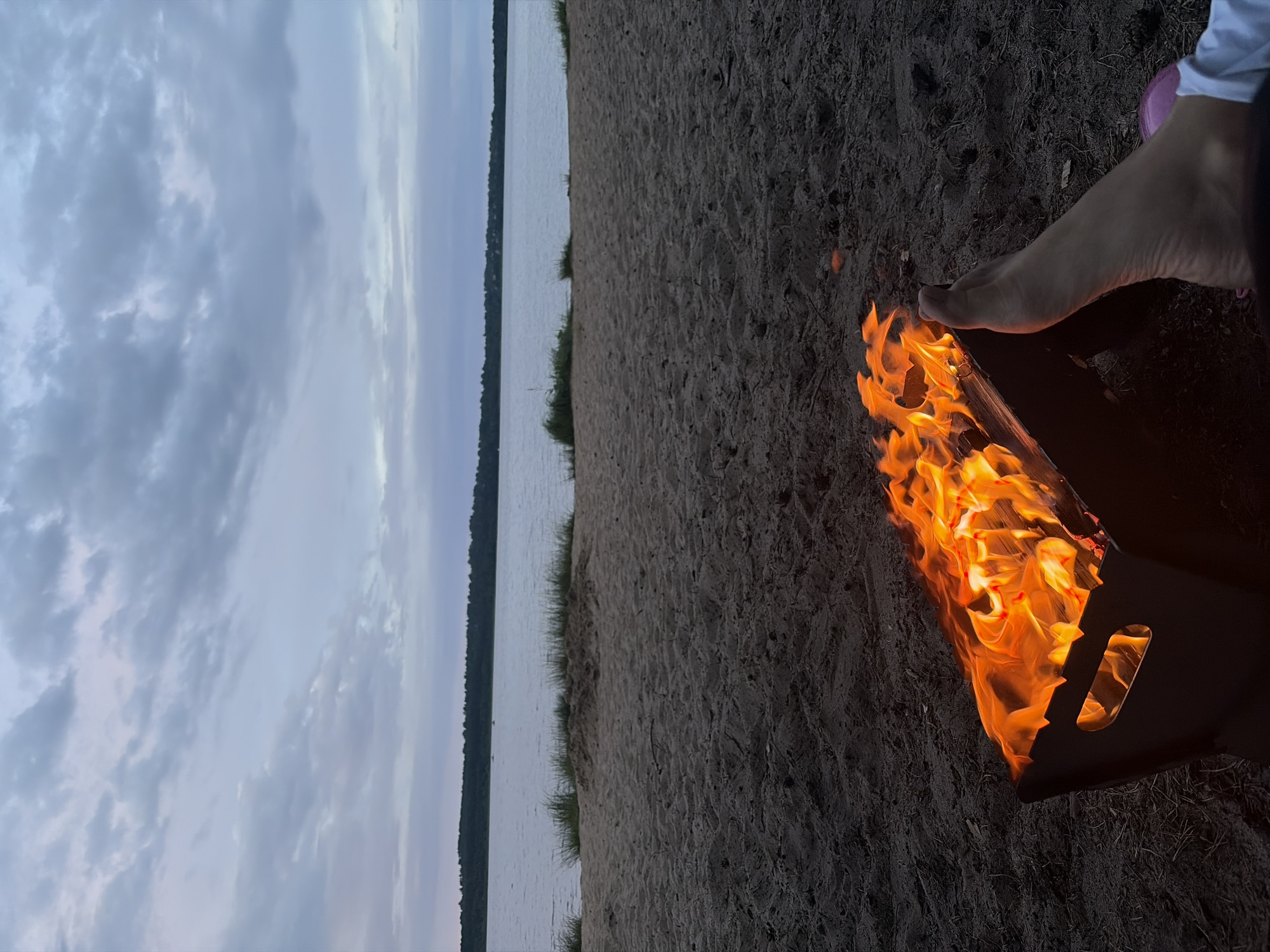 This is a medium shot of a beach scene at dusk, with a fire burning in a portable fire pit in the foreground. A person's foot is visible to the right of the fire pit. In the background, a body of water stretches to the horizon, with a tree-lined shore and a cloudy sky above. The sand is dark and textured, with some dried grass and debris scattered on it. The fire is bright orange and yellow, with visible flames and glowing embers. The water is calm and reflects the muted colors of the sky. The overall atmosphere is peaceful and serene. The location is the shore of a lake in Vaskelovo, Russia.
