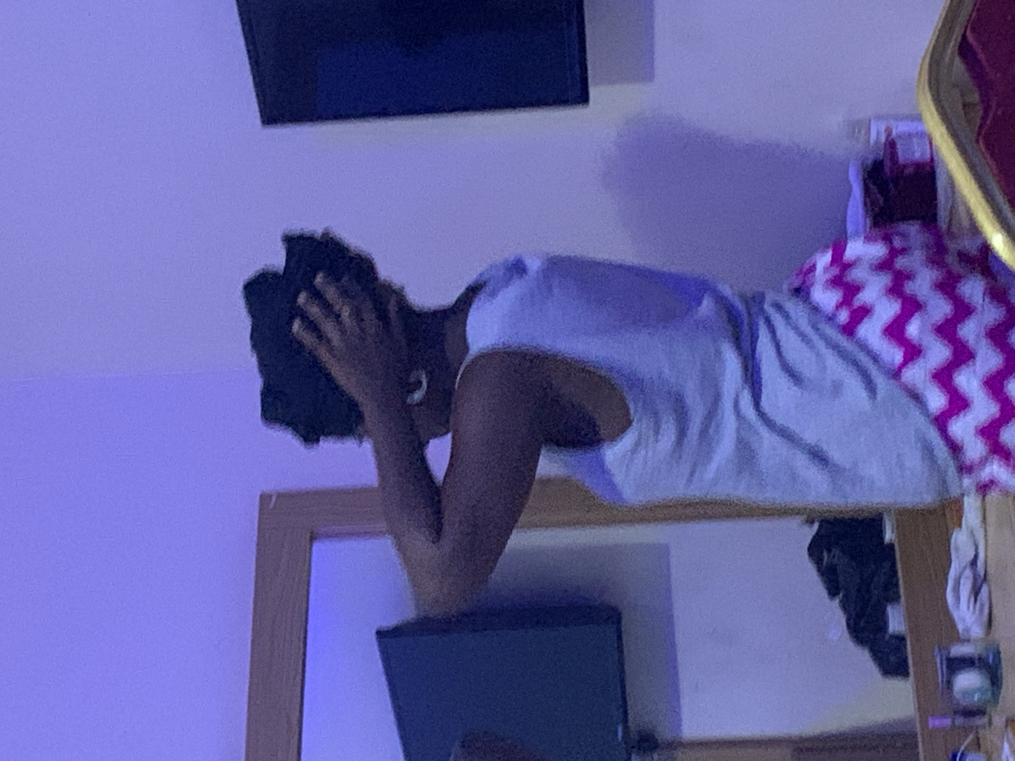 A person is lying on what appears to be a bed or surface in a room. The person has their hands on their head, possibly indicating distress or reflection. A mirror is visible, reflecting a television set. A bedspread with a pink and white zig-zag pattern is present. The environment has a bluish-purple lighting. No contextual information suggests this photo was taken in Lokoja, Nigeria.
