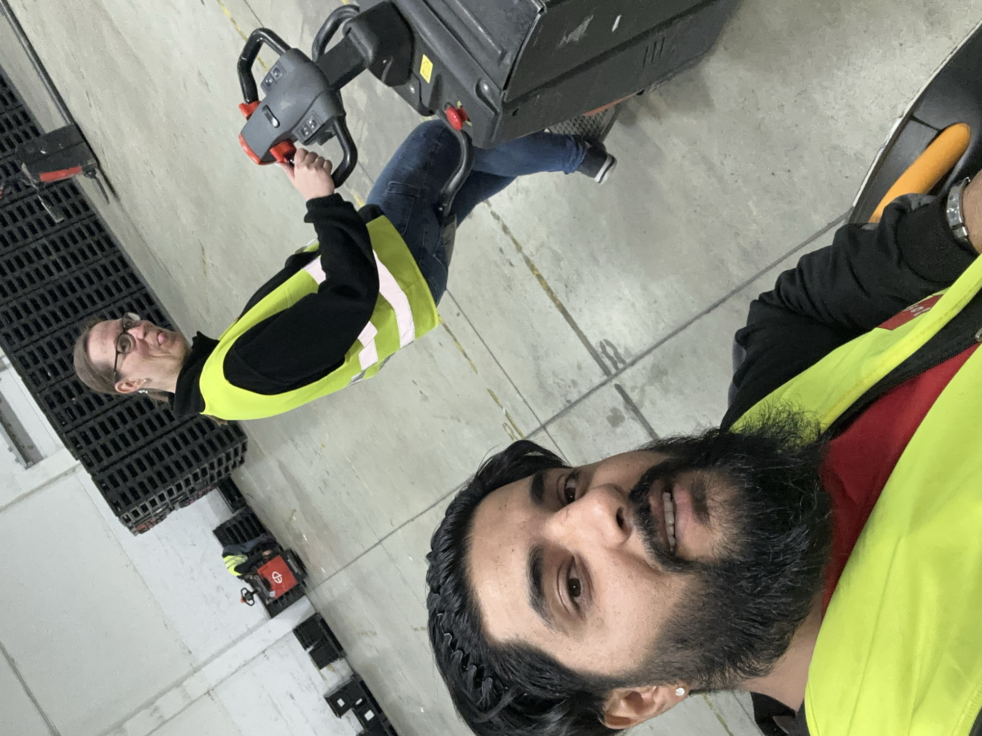In an indoor warehouse environment, likely in Schweitenkirchen, Germany, two individuals are present. In the foreground, a man with dark hair and a beard, wearing a bright yellow safety vest over a red shirt, is taking a selfie. He is positioned on a black pallet jack. In the mid-ground, a person with glasses, also wearing a yellow safety vest, operates another pallet jack. This pallet jack is black and red. The background features tall stacks of black pallets and a white wall. The floor is concrete with yellow painted lines.