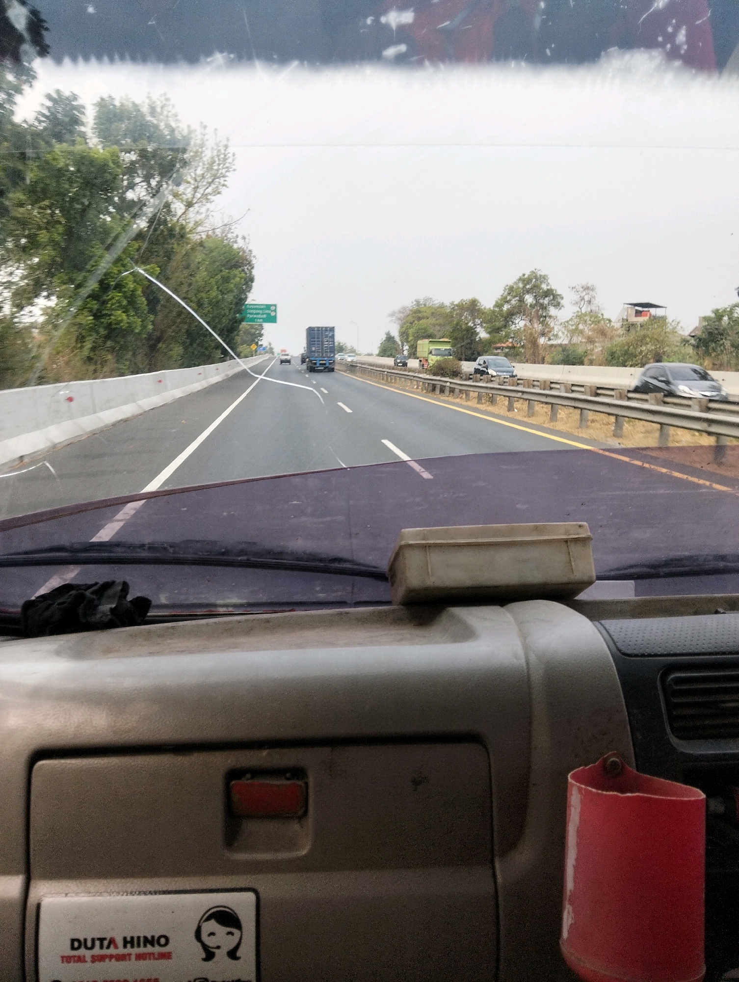 The image is a point-of-view shot from inside a vehicle on a highway, likely in or near Semarang, Indonesia. The windshield of the vehicle has a prominent crack running across it. Through the windshield, a multi-lane highway stretches ahead under an overcast sky. Traffic is visible on the highway, including cars and trucks. On the left side of the highway, there is a concrete barrier and vegetation. On the right side, there is a metal guardrail and more vegetation. The interior of the vehicle is visible in the foreground, showing a dashboard with a red cup holder on the right and a sticker that reads "DUTA HINO TOTAL SUPPORT HOTLINE" on the left.