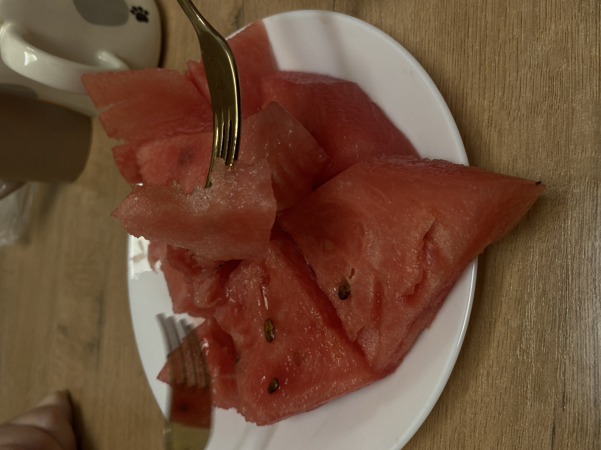 In Votkinsk, Russia, a white plate filled with sliced watermelon is positioned on a wooden table. A golden fork is used to pick up a piece of watermelon. In the background, two mugs are visible, one brown and one white with a gray elephant design and a paw print. A person's hand is partially visible in the upper left corner of the image. The scene captures a moment of enjoying fruit.