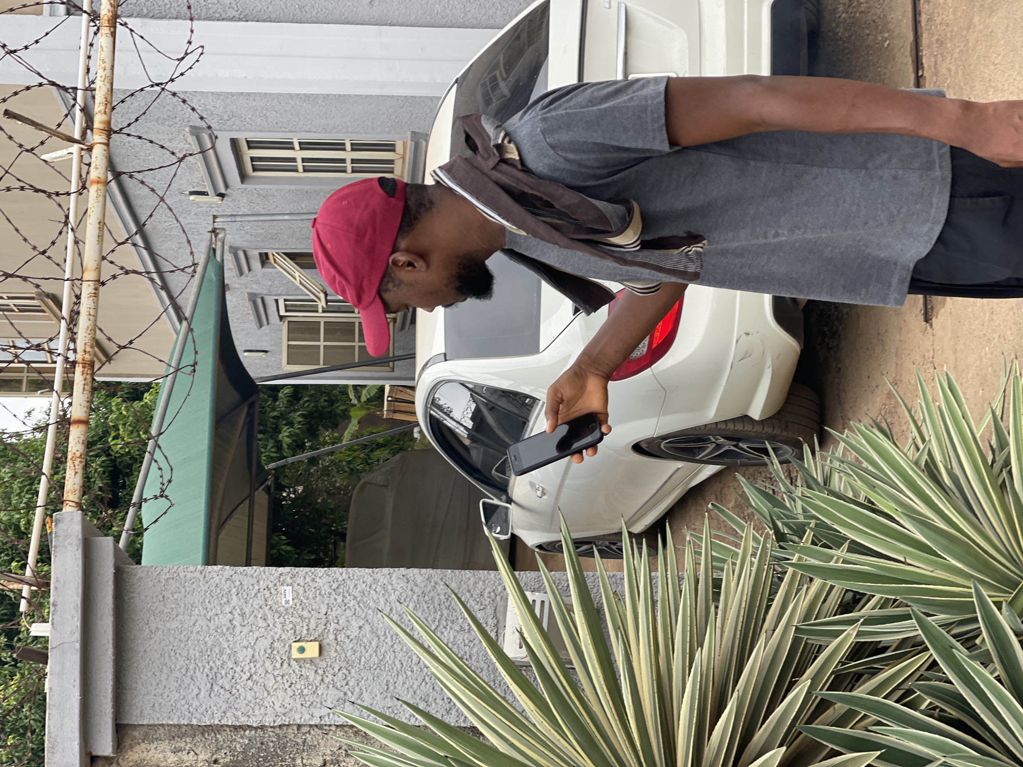 The image, taken in Abuja, Nigeria, depicts a man standing outdoors, engaged with his smartphone, during bright daylight hours. The man, viewed in profile, wears a red baseball cap, a grey short-sleeved t-shirt, and dark shorts. A brown and beige striped scarf is draped around his neck. He has a beard and is holding a black smartphone in his right hand, looking down at its screen.
Behind the man, a white sedan is parked, with its rear section visible. The background includes a grey textured building featuring multiple windows and a green awning or canopy. To the far left, a rusty metal pole wrapped with barbed wire suggests a boundary. The foreground and the right side of the frame are filled with spiky plants characterized by green and white variegated leaves. A grey textured wall or fence is also prominent in the foreground, with a small, indistinct white label and a small yellow-green rectangular object (possibly a doorbell) attached to it. The overall scene conveys a casual, everyday moment in a residential or suburban setting.