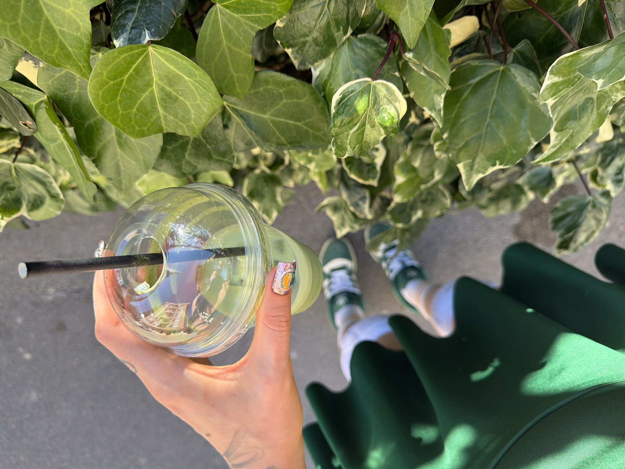 A high-angle, eye-level shot shows a person holding a clear plastic cup with a dark straw. The cup appears to contain a beverage. The person's hand is visible, with a decorated nail on the thumb. The person is wearing a green dress and green and white sneakers. The background consists of green ivy leaves. The reflection in the cup indicates the setting to be the city of Budva, Montenegro.