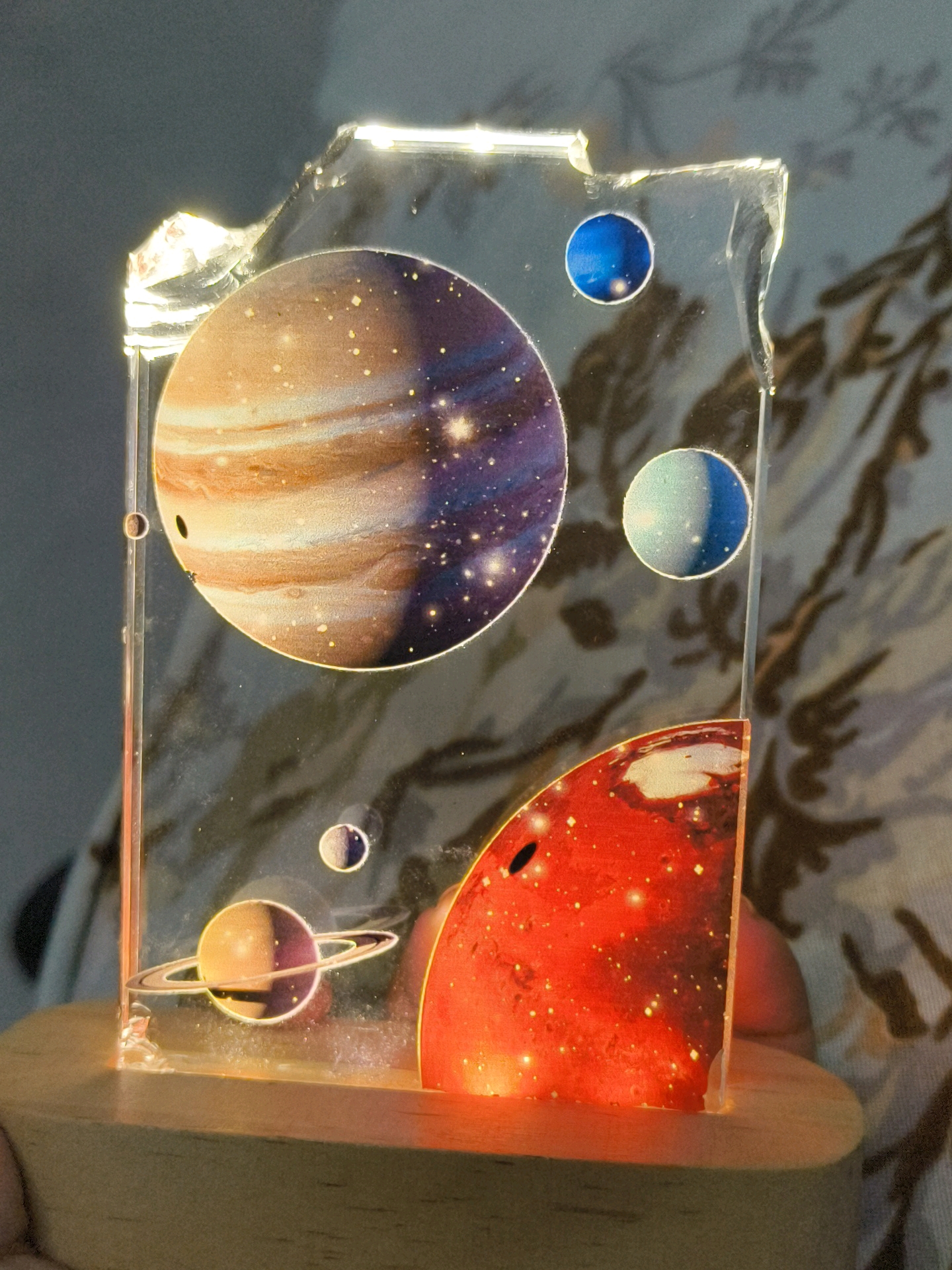 A close-up, vertical, eye-level shot showcases an acrylic night light shaped like a solar system. The acrylic panel is imprinted with images of planets and stars, and is lit from behind by embedded LEDs. The panel is designed with a jagged top edge, showing imperfections where it has been cut or broken. At the bottom, the acrylic piece rests on a light-colored wooden base. The background consists of a patterned fabric, likely a blanket or curtain, with dark, stylized floral or leaf designs. Several planets are visible: a large, swirling gas giant with prominent bands and a dark spot dominates the upper left quadrant. Smaller, spherical planets in shades of blue and green are scattered in the upper right. A ringed planet, resembling Saturn, is positioned in the lower left, with a smaller moon nearby. A bright, reddish-orange planet, possibly Mars, occupies the lower right section. The lighting from the night light casts a warm glow on the wooden base and illuminates the acrylic, creating a starry effect within the depicted planets. The image is sharp and clear, focusing on the night light. There are no people visible in the image. The location is unknown.