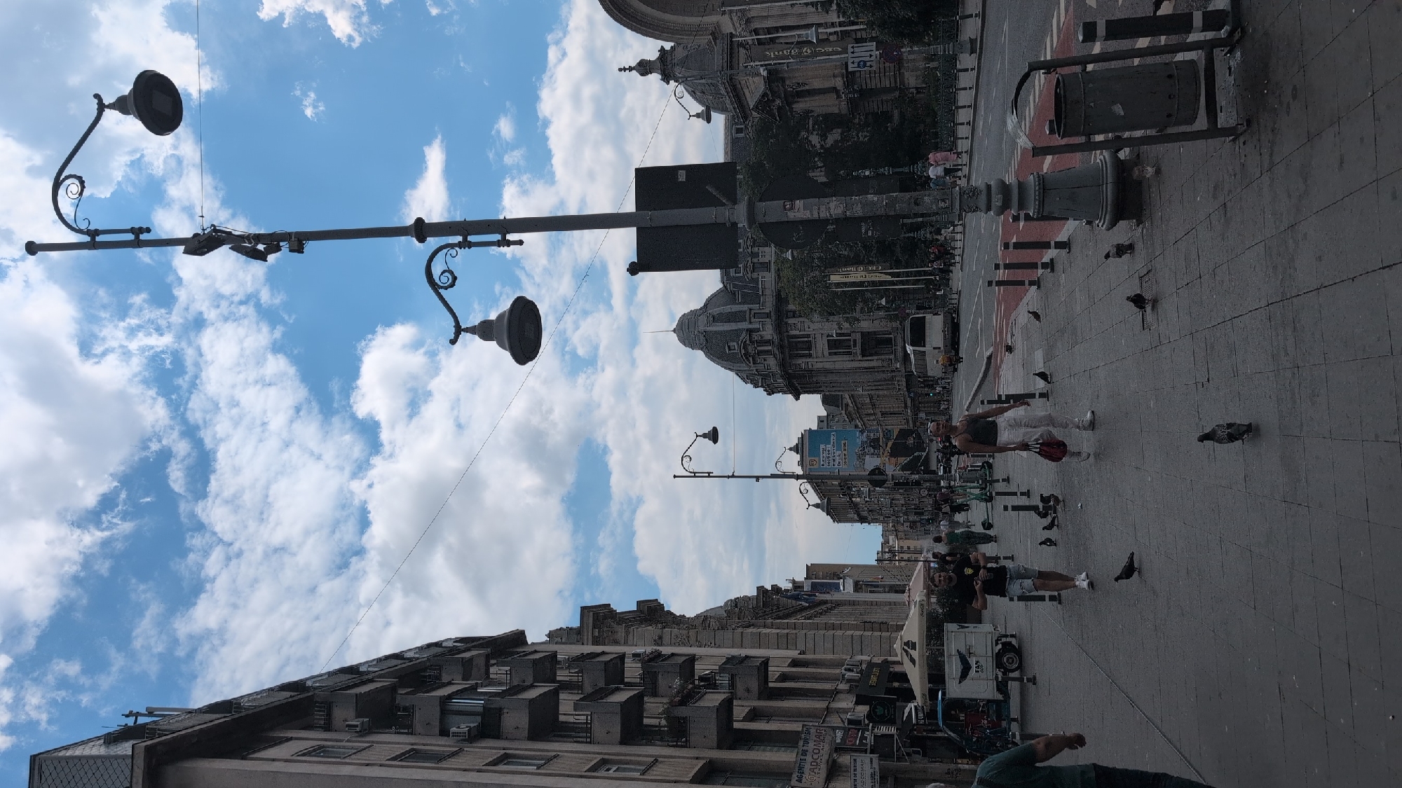 The image captures a view down a wide city street in Bucharest, Romania, under a sky with scattered white clouds. The street is lined with imposing, multi-story buildings featuring balconies and ornate architectural details. A prominent streetlamp with multiple fixtures stands tall on the right side of the frame, casting long shadows.
On the sidewalk in the foreground, several people are walking, and a number of pigeons are scattered across the pavement, some pecking at the ground. A man in a dark t-shirt and shorts is walking with his head down, while a person in white pants and a dark tank top walks towards the right, carrying a red bag. Further down the sidewalk, more pedestrians can be seen.
The street itself has a bus lane marked with red paint, and a white bus is visible in the distance. Various street furniture, including bollards, a trash can, and more streetlights, are present. A small vendor cart with umbrellas is visible on the left side of the sidewalk. The overall impression is of a bustling urban environment on a partly cloudy day.