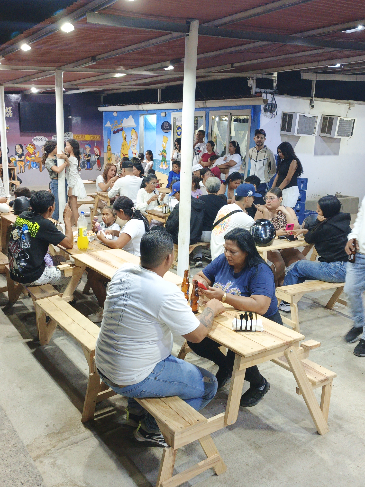 The image shows a bustling outdoor dining area in Villa Marina, Venezuela, likely in the evening. The setting features wooden picnic tables and benches occupied by numerous people, indicating a social gathering or restaurant scene. Individuals are seen sitting, talking, and using their phones. Children are also present. The area is sheltered by a roof supported by white pillars. A mural depicting cartoon characters and other figures adorns the wall. Air conditioning units are visible on a white building section, suggesting a warm climate. Beer bottles are placed on tables, and some people are holding them. The floor is concrete.