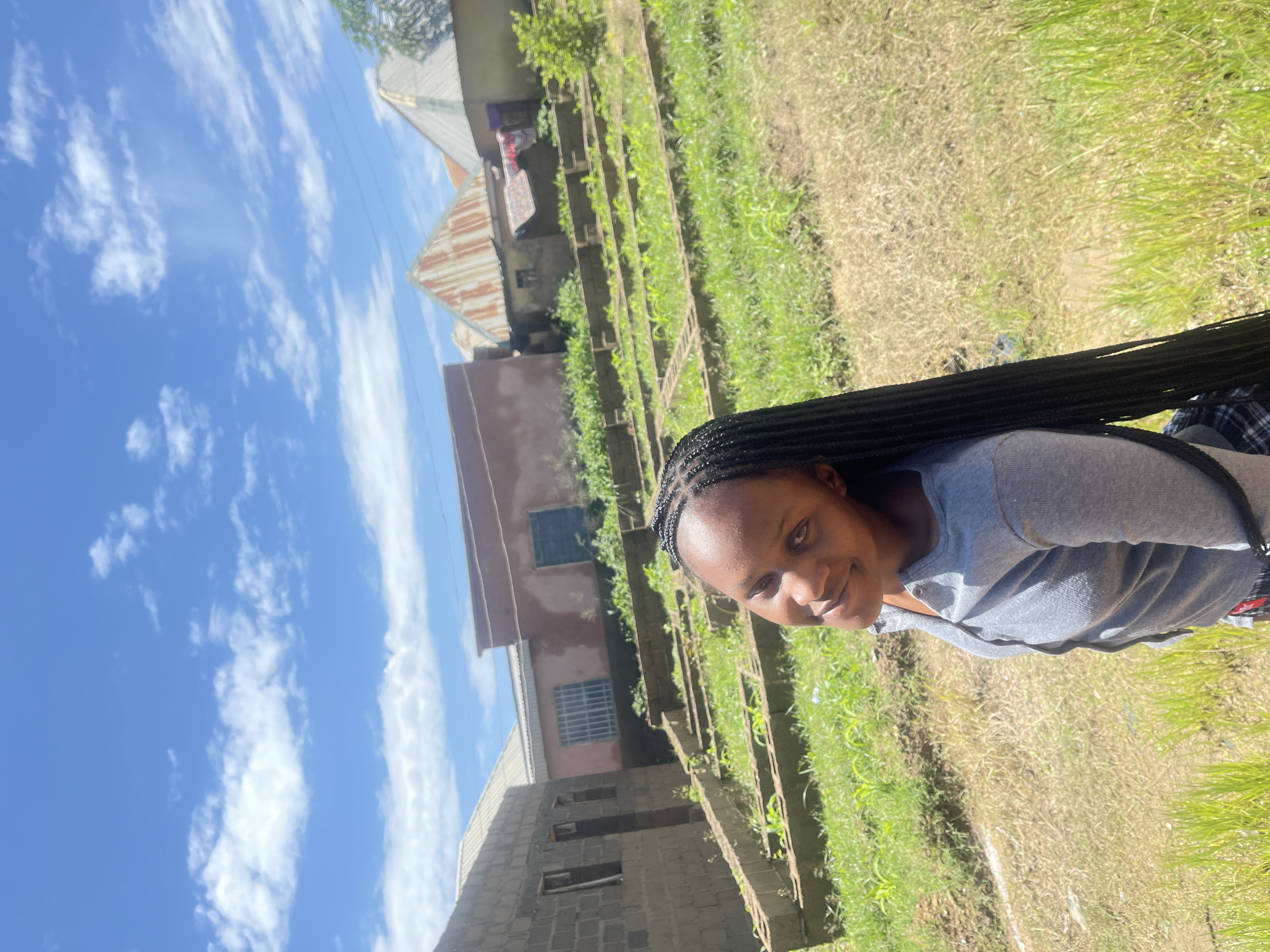 In Rafin Par, Nigeria, a young Black woman with long, braided hair smiles at the camera in a selfie. She is wearing a grey long-sleeved shirt. The background shows a rural or semi-rural setting with several unfinished buildings constructed of concrete blocks. In front of the buildings are rows of raised garden beds with green plants, likely young crops. The sky is blue with scattered white clouds. The overall scene suggests a sunny day in a developing area.