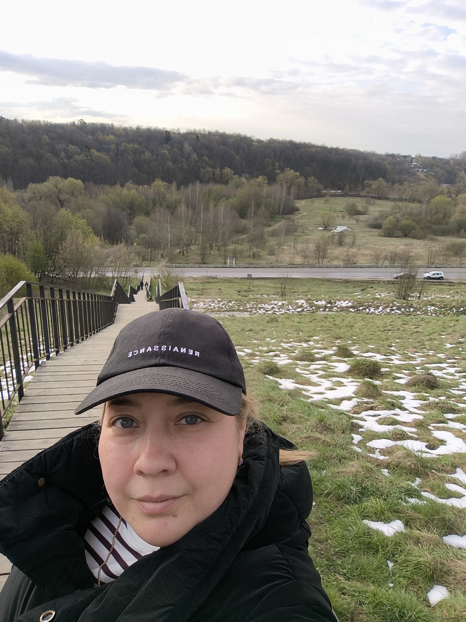 A selfie shows a woman wearing a black "Renaissance" baseball cap and a black winter coat, with a striped shirt and a gold chain necklace visible at the collar. She is standing at the top of a wooden staircase with black metal railings. The staircase leads down to a grassy area with patches of snow. Beyond the grassy area, there is a road with a white car visible. In the background, there is a forest. The photograph was taken in the Vorobyovy Gory District of Moscow, Russia.