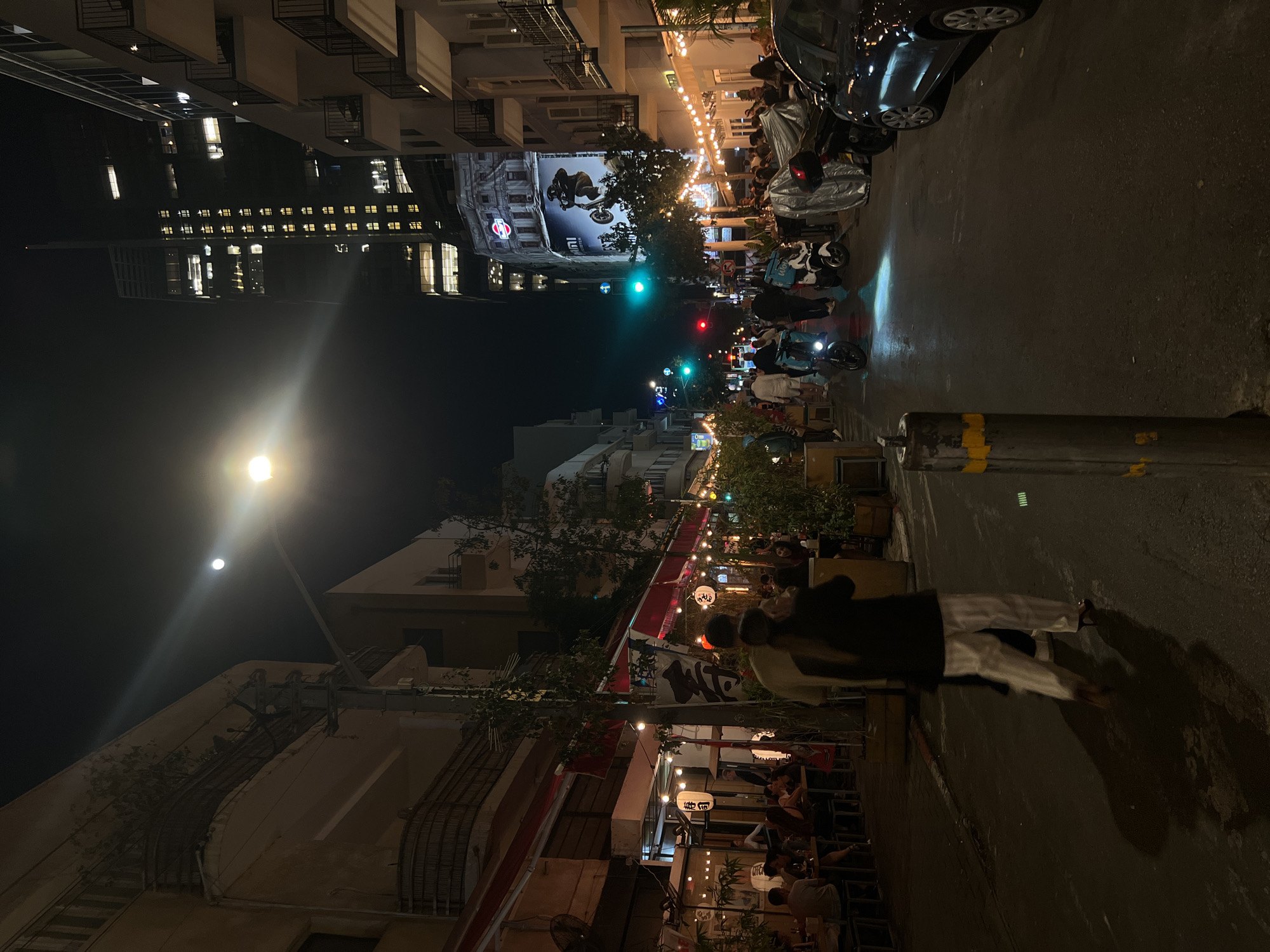 The image shows a night scene on a street in Tel Aviv, Israel. The street is crowded with people, some walking, others sitting at tables outside restaurants and bars, which are decorated with string lights and Japanese lanterns. Motor scooters and cars are parked on the side of the road. Traffic lights are visible in the distance. High-rise buildings loom in the background. Two people are walking on the right side of the street, their backs to the camera. A lamppost stands on the left, casting a bright light.