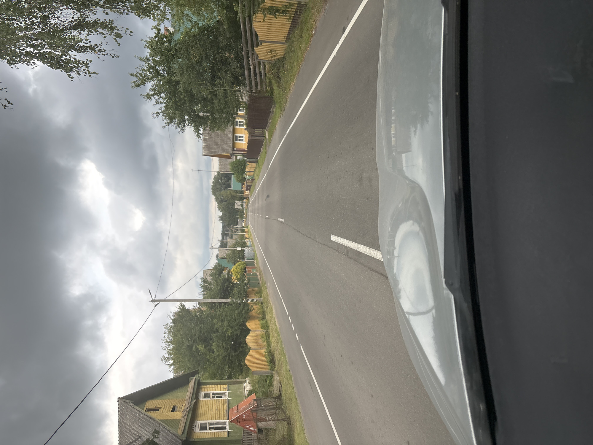 The image depicts a street scene in the village of Usełub, Usełubski Selski Sovet, Belarus, under an overcast sky. The perspective is from the interior of a vehicle, looking out through the windshield at a paved road stretching into the distance. The road is lined with houses and fences, characteristic of a rural Belarusian settlement. On the left side of the road, a yellow wooden house with white trim and dark green shutters is visible, along with a small red structure and a rustic wooden fence. On the right, a series of wooden fences, some painted yellow and others a natural wood color, separate properties from the road. Further down the street, more houses with varied architectural styles and colors can be seen, interspersed with trees and vegetation. Utility poles and wires are present along the roadside. The overall atmosphere is calm and ordinary, with no people or notable activity captured in the frame. The road itself features white dividing lines, indicating it is a two-lane road. The foreground is dominated by the car's dashboard and windshield.