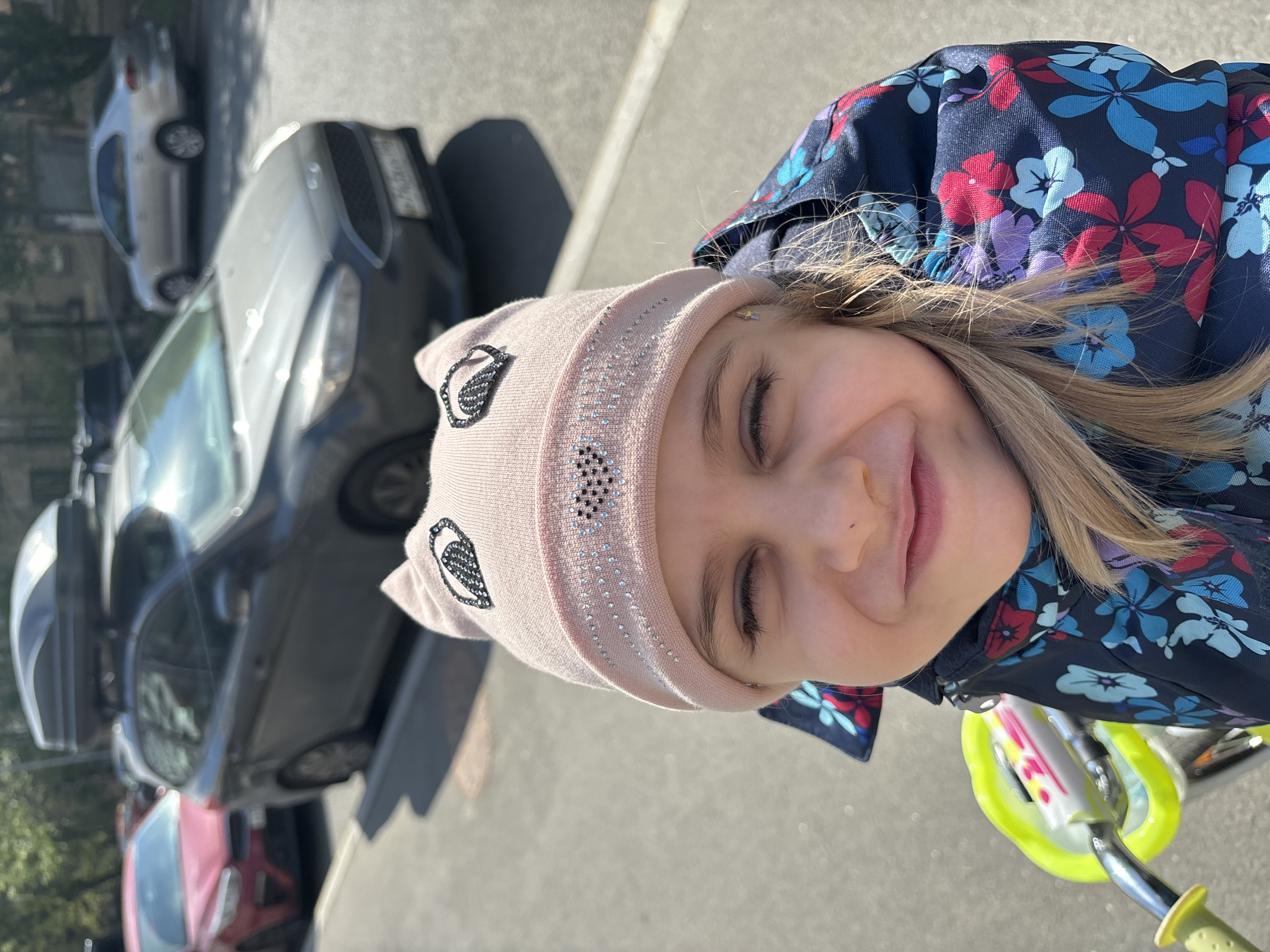 A close-up, slightly angled, outdoor shot depicts a young girl smiling with her eyes closed. She is wearing a light pink hat with cat ear details and floral accents, along with a navy blue jacket featuring a floral pattern. The girl is positioned on or near a lime green tricycle.
The background includes several parked cars, one of which is gray and another which is red. The setting appears to be a street in an urban area. There is no evidence in the image to support the claim that the location is Saint Petersburg, Russia.