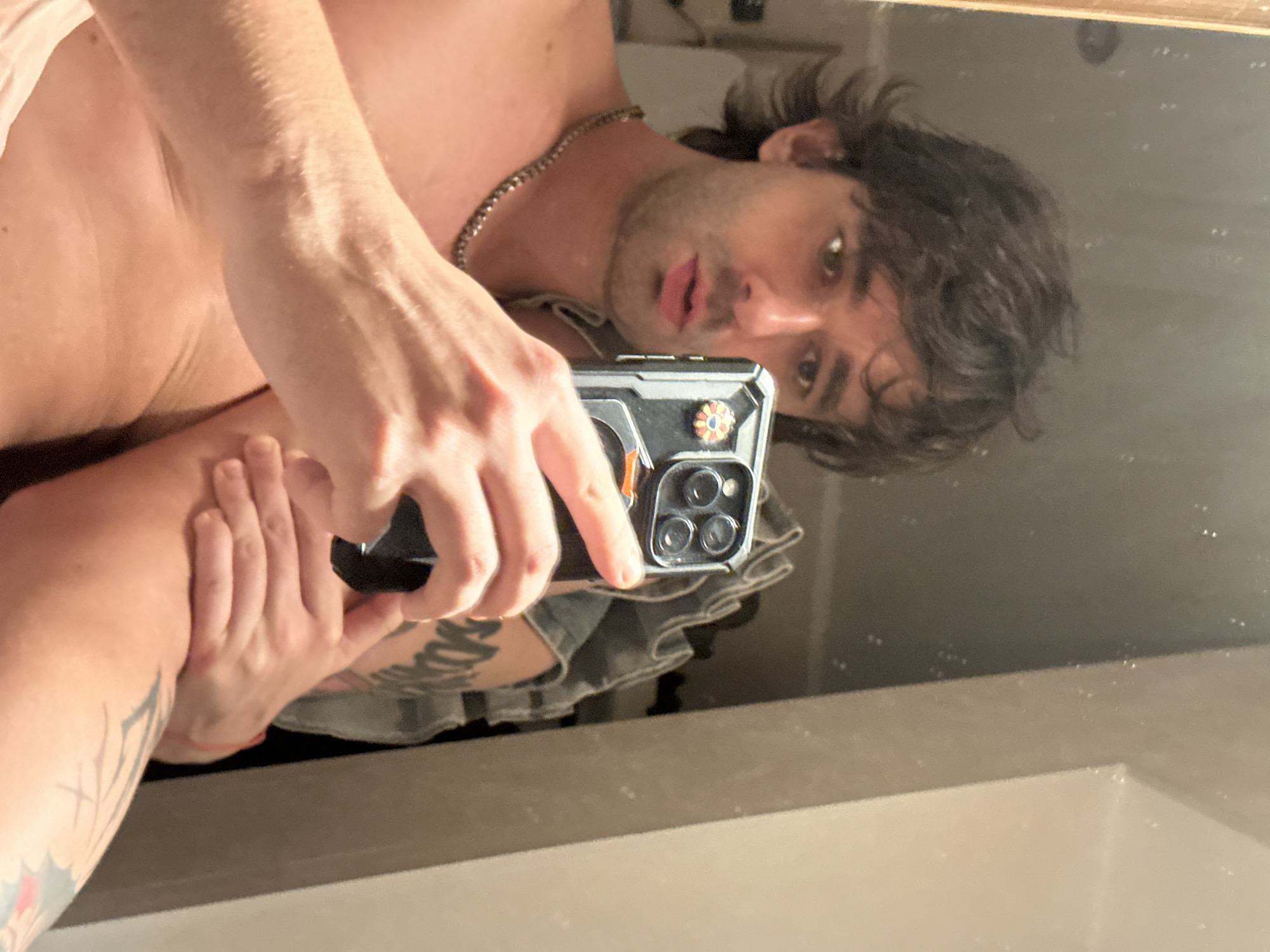 A person is taking a mirror selfie in a bathroom. The person has brown, wavy hair and is wearing a necklace. They are holding a smartphone in their right hand, which is angled towards the mirror. The background shows a tiled wall and a reflection of another person, possibly also a selfie. The overall lighting is dim, suggesting the photo was taken indoors.