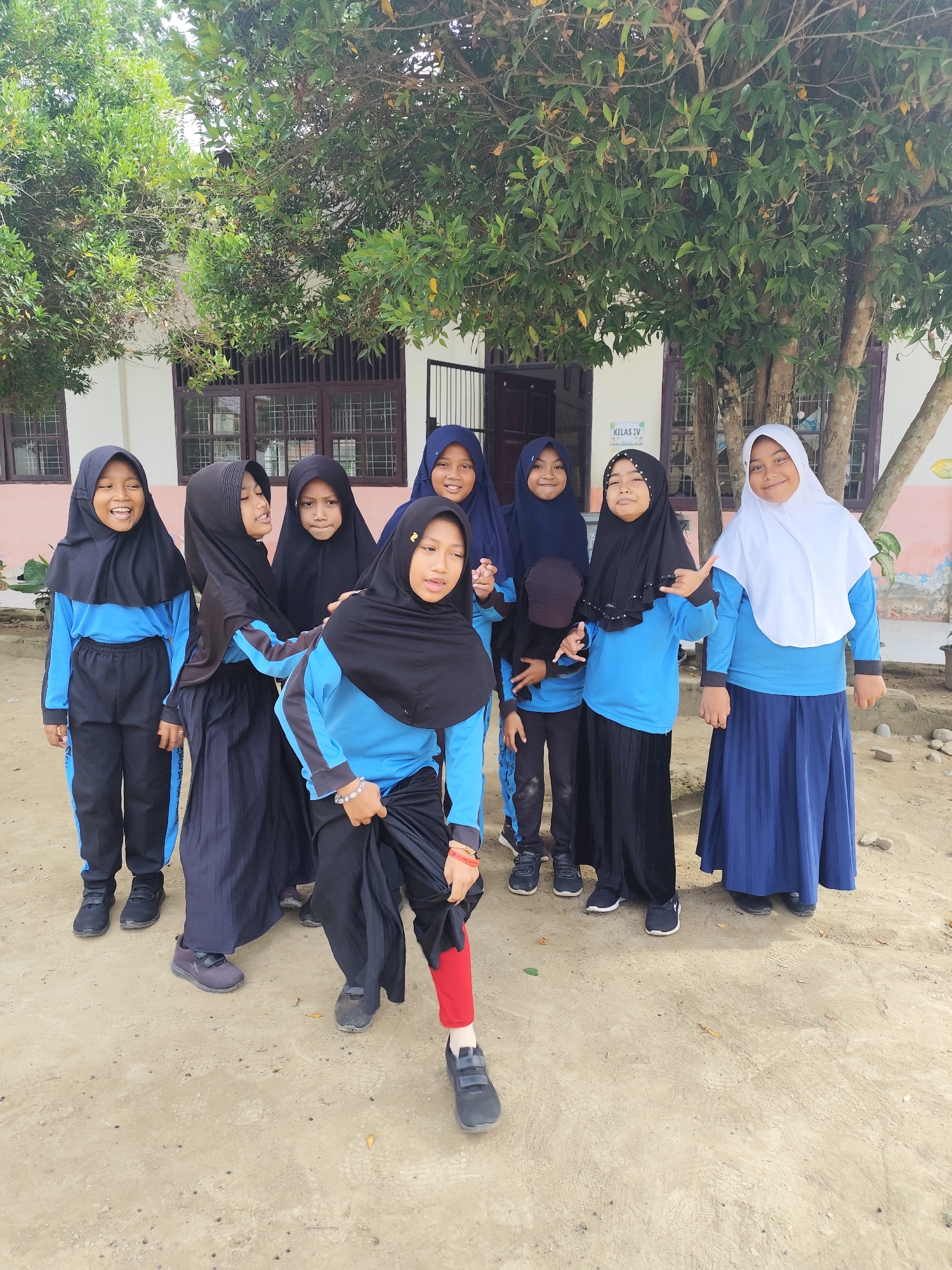 A group of eight young girls, all wearing hijabs and school uniforms consisting of blue tops and black pants or skirts, are posing for a photo outdoors. They are standing in front of a building with windows and a tree with green foliage. The ground beneath them is dirt.
The girl on the far left is laughing with her mouth open. The girl next to her is also smiling and looking at the camera. The next three girls are clustered together, with the middle one in black looking off to the side. The girl in the bright blue shirt is striking a pose with her hand up. The girl next to her is wearing a white hijab and a blue skirt and is smiling at the camera. The last girl in the back is wearing a dark blue hijab and smiling.
The setting appears to be a schoolyard. The lighting suggests it is daytime, likely late morning or early afternoon, with bright, diffused light. The sky is not visible.
There is a sign visible on the building behind the girls that reads "KELAS IV," which translates to "CLASS IV" in Indonesian. The overall mood seems cheerful and energetic, with the girls smiling and interacting with each other.
