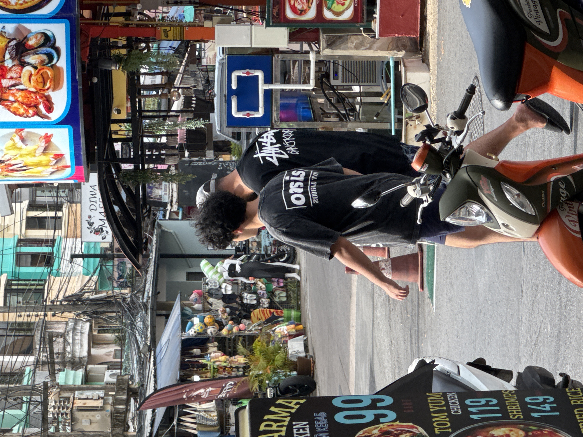 The image captures a street scene in Karon, Thailand. In the foreground, two individuals are visible. The person on the left is wearing a dark t-shirt with "OASIS" printed on the back and dark shorts. The person on the right is wearing a dark t-shirt with "STUSSY BANGKOK" printed on it and dark shorts. Both are walking away from the camera. A motorcycle is parked in the lower left quadrant of the image, and another motorcycle is partially visible on the right.
Behind the individuals, a market stall is set up with various items displayed, including bags, clothing, and accessories. Further back, buildings with colorful facades and intricate wiring dominate the urban landscape. A sign with food prices, "TOM YUM CHICKEN 119 SHRIPS 149 FRIED RICE," is prominently displayed on a stand in the lower left. A blue sign with a white graphic resembling a simplified television set or a control panel is mounted on a structure in the middle ground. The overall environment suggests a busy commercial area with a vibrant street market. The lighting indicates it is daytime, possibly morning or afternoon.