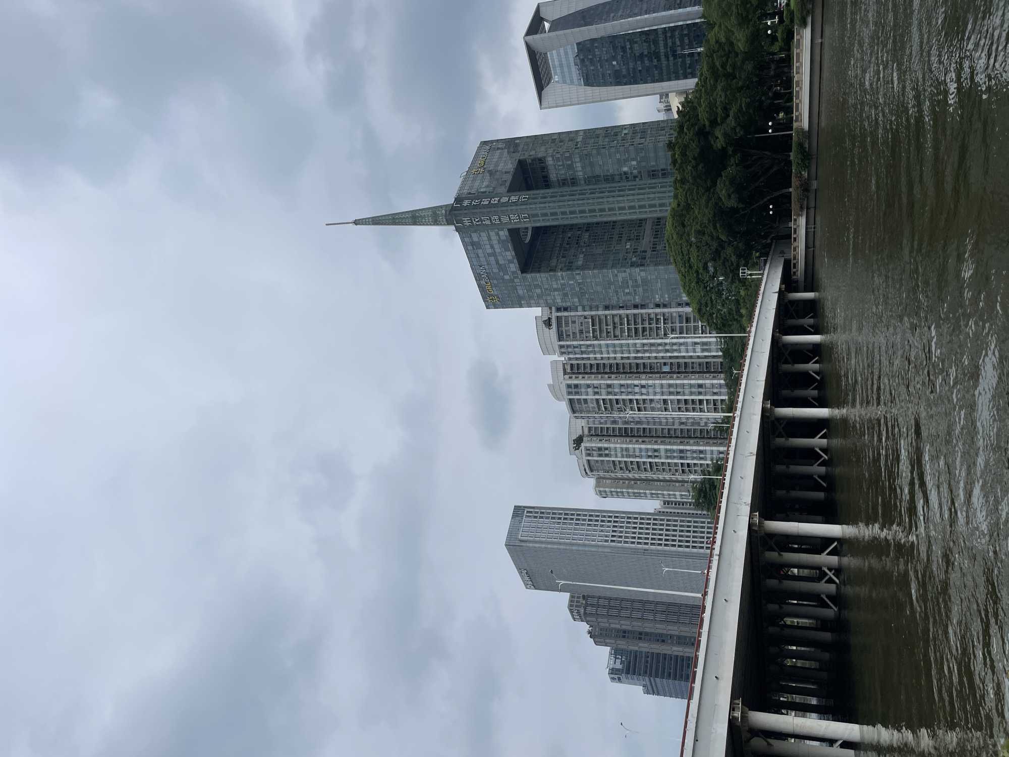 The photograph, taken in the city of Tianhe District, China, shows a waterfront scene under an overcast sky. The view is from a bridge overlooking the river. The photograph contains several skyscrapers and office buildings. The skyline features modern architecture, including a prominent structure with a central opening and a spire. The building has the words "GAC BANK" on the upper floors. Along the river, there is a tree-lined park. The water of the river has dark green color. The bridge's structure is visible with white support pillars extending into the water.