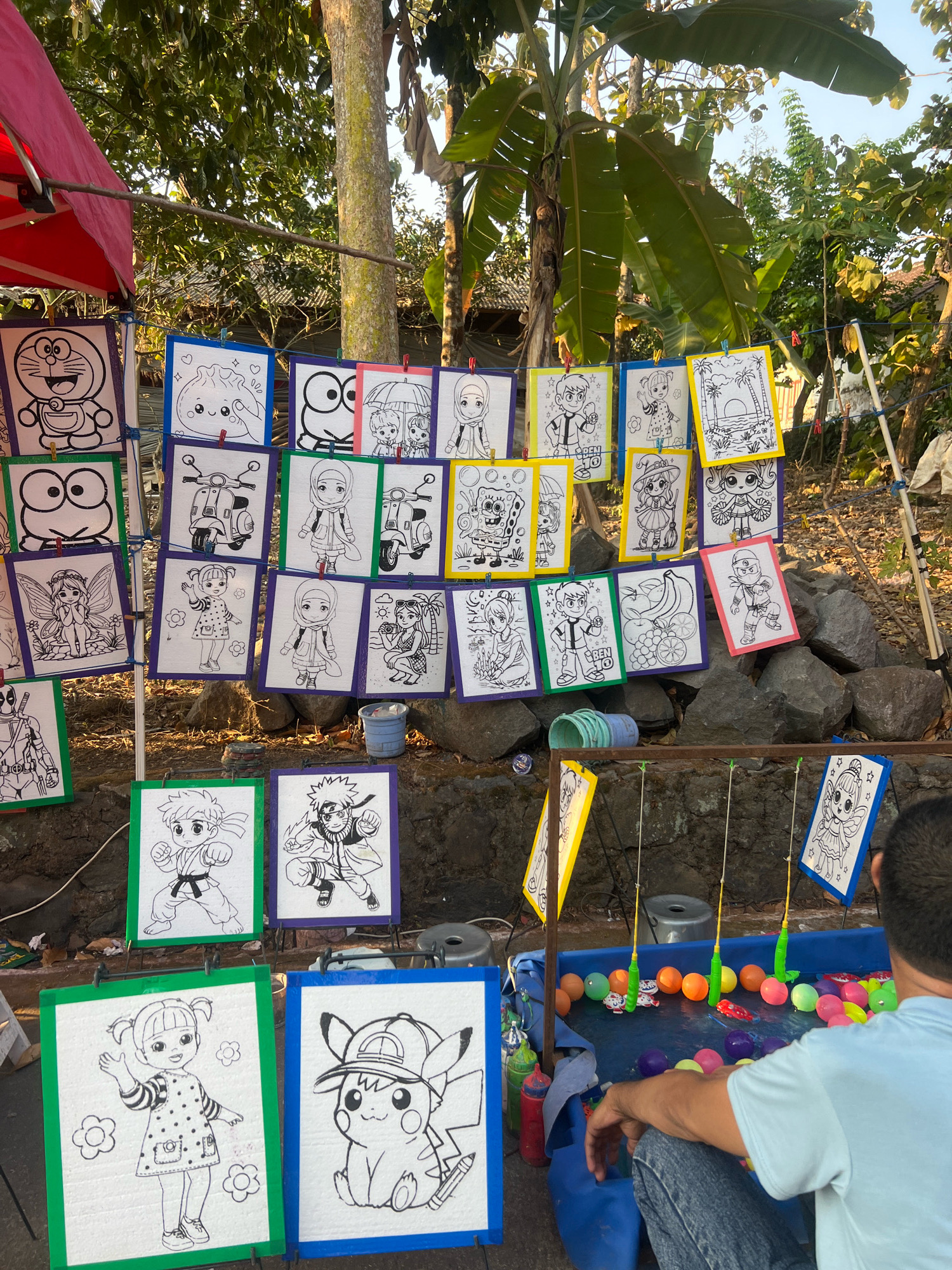The image depicts an outdoor stall, possibly in the city of Madyocondro, Indonesia, featuring an array of coloring pages. These pages, mounted on various colored boards (pink, blue, green, yellow, and white), showcase a variety of cartoon characters, including Doraemon, Spongebob Squarepants, Hello Kitty, and characters resembling anime figures. Some pages also feature everyday objects like a scooter, a fairy, a young girl, a sunset landscape, and a fruit still life. The coloring pages are suspended by clips or ties on a string or wire, forming a colorful display against a backdrop of trees and rocks.
In the foreground, a blue plastic pool contains colorful balls and small fishing rods, suggesting a game of chance or skill. A person, seen from behind with their back to the camera, is seated near the pool, looking towards it. They are wearing a light blue t-shirt and denim jeans. The overall scene is informal and likely part of a local market or fair. The lighting suggests it is daytime, with sunlight casting shadows and illuminating the subjects. The environment appears somewhat rustic, with visible rocks and natural foliage.
