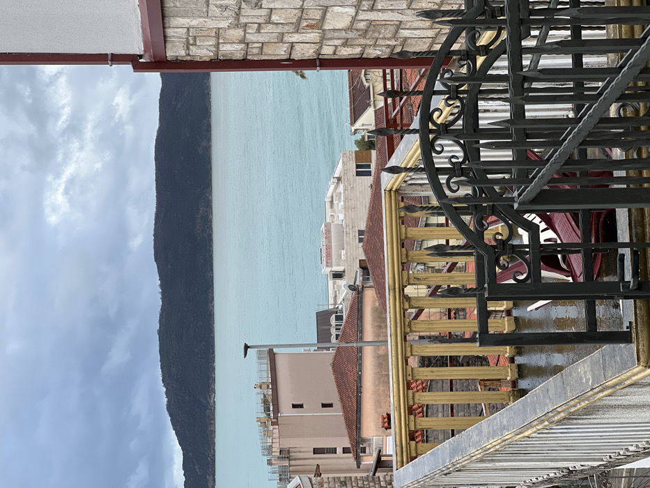 This image presents an elevated, static view over a residential area of Budva, Montenegro, overlooking the Adriatic Sea under an overcast sky.
In the immediate foreground, a wet balcony or terrace surface is visible, bounded by a white and grey railing on the left. Adjacent to this, a golden-yellow balustrade composed of columnar supports runs horizontally. To the right, a dark grey wrought iron gate, featuring ornate scrolled details and pointed finials, stands partially open or positioned. Behind this gate, two red chairs are overturned or collapsed on the wet ground.
The midground reveals a dense arrangement of building rooftops and structures. Multiple buildings exhibit terracotta-tiled roofs, interspersed with flat-roofed edifices. One distinct, multi-story building features a light brown-pink facade. Another light-colored, flat-roofed building displays the Cyrillic text "ПРОДАЈА ЗАЛЕ," which translates to "SALE," indicating a property for sale. A tall, dark grey street lamp or utility pole extends vertically from this midground cluster.
The background consists of the turquoise-hued Adriatic Sea, which extends horizontally across the frame. Beyond the sea, a large, dark green, forested landmass, consistent with an island or peninsula, occupies the distant horizon. The sky above is entirely overcast with heavy grey clouds. No individuals, animals, or explicit dynamic actions are observed within the scene.
