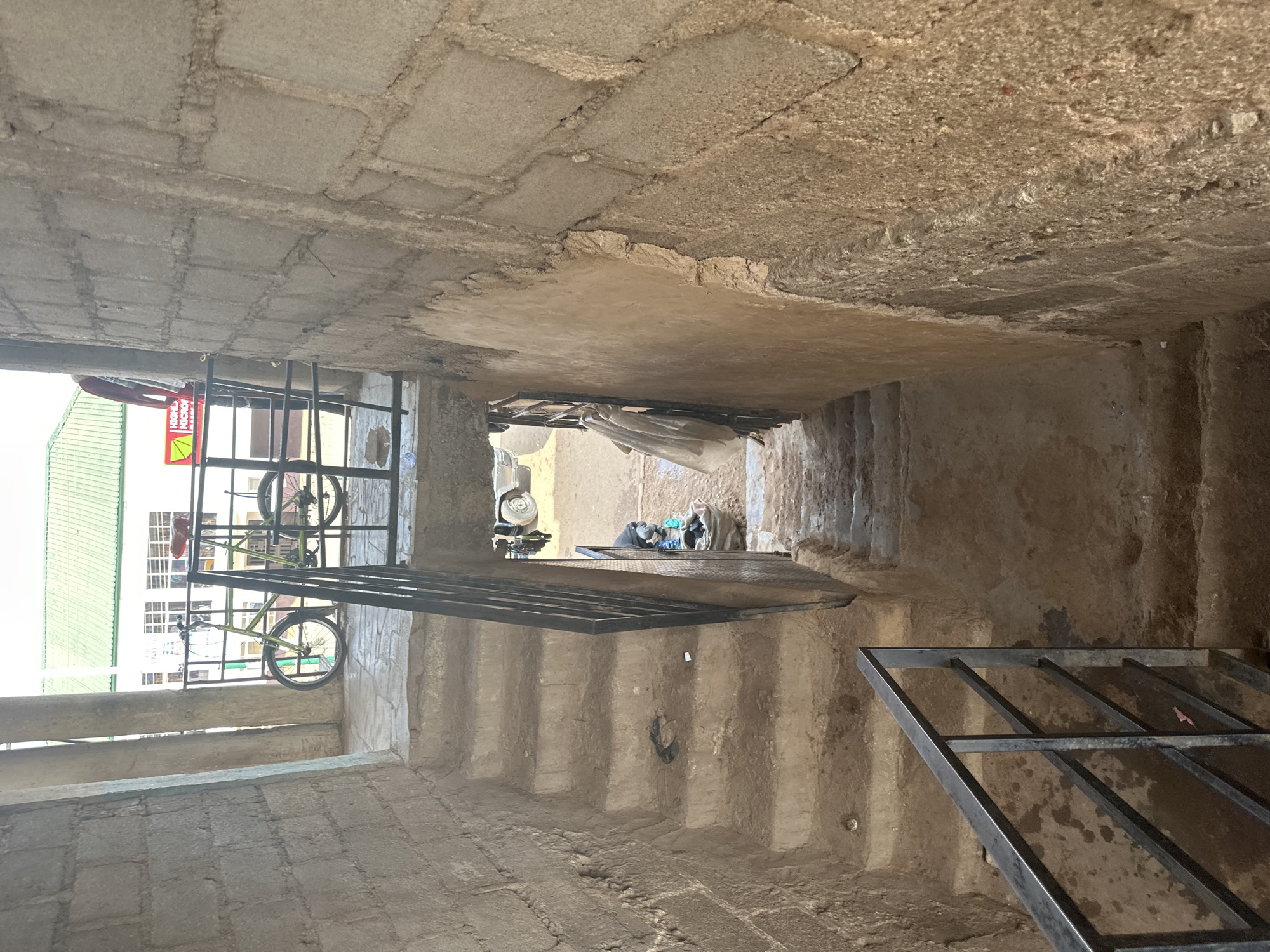 This is a street-level, medium shot of an outdoor staircase in Bukuru, Nigeria. The staircase is made of concrete with rough, unplastered stone walls on either side. The stairs descend from a landing where a bicycle is parked near a metal railing. In the background, a building with a green roof and white windows is visible. A portion of a red and yellow sign with "HIGHLY" and "MICRO" visible is attached to the building. A person is partially visible on the landing, wearing a red shirt and leaning against a railing. Further down the stairs, a person is seen near a large sack, possibly sorting items. A light-colored vehicle is parked in the background at the bottom of the stairs. The overall scene depicts a functional, somewhat utilitarian outdoor space.