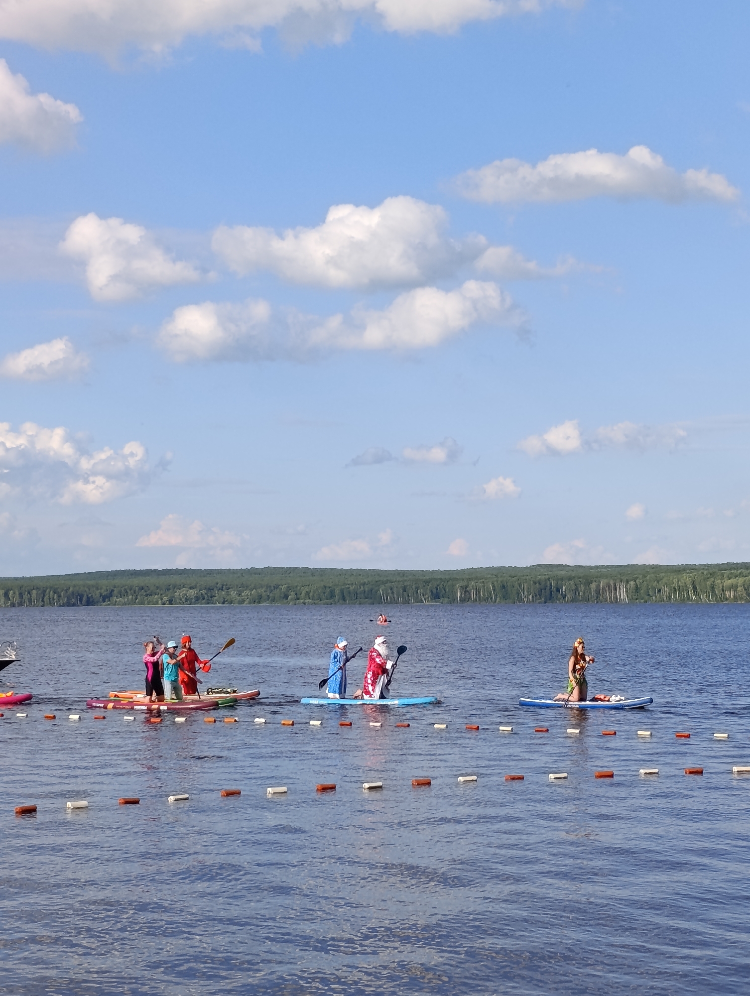 A group of people are paddleboarding and kayaking on a wide, calm lake under a bright blue sky with fluffy white clouds. The scene appears to be a festive event, as several participants are dressed in costumes. One person on a paddleboard is dressed as Santa Claus, complete with a red suit and hat, and is holding a paddle. Another person on a paddleboard is wearing a blue Santa costume. Two people are in a kayak, one dressed in pink and another in a bright orange outfit. Further out on the lake, another person on a paddleboard is wearing a floral skirt and lei, seemingly dressed for a luau. The lake is lined with buoys, suggesting a designated area for water activities or a race. In the background, a dense forest with green trees stretches across the horizon. The time of day appears to be midday, with clear, sunny weather. The atmosphere seems joyful and recreational. No visible text is present in the image.