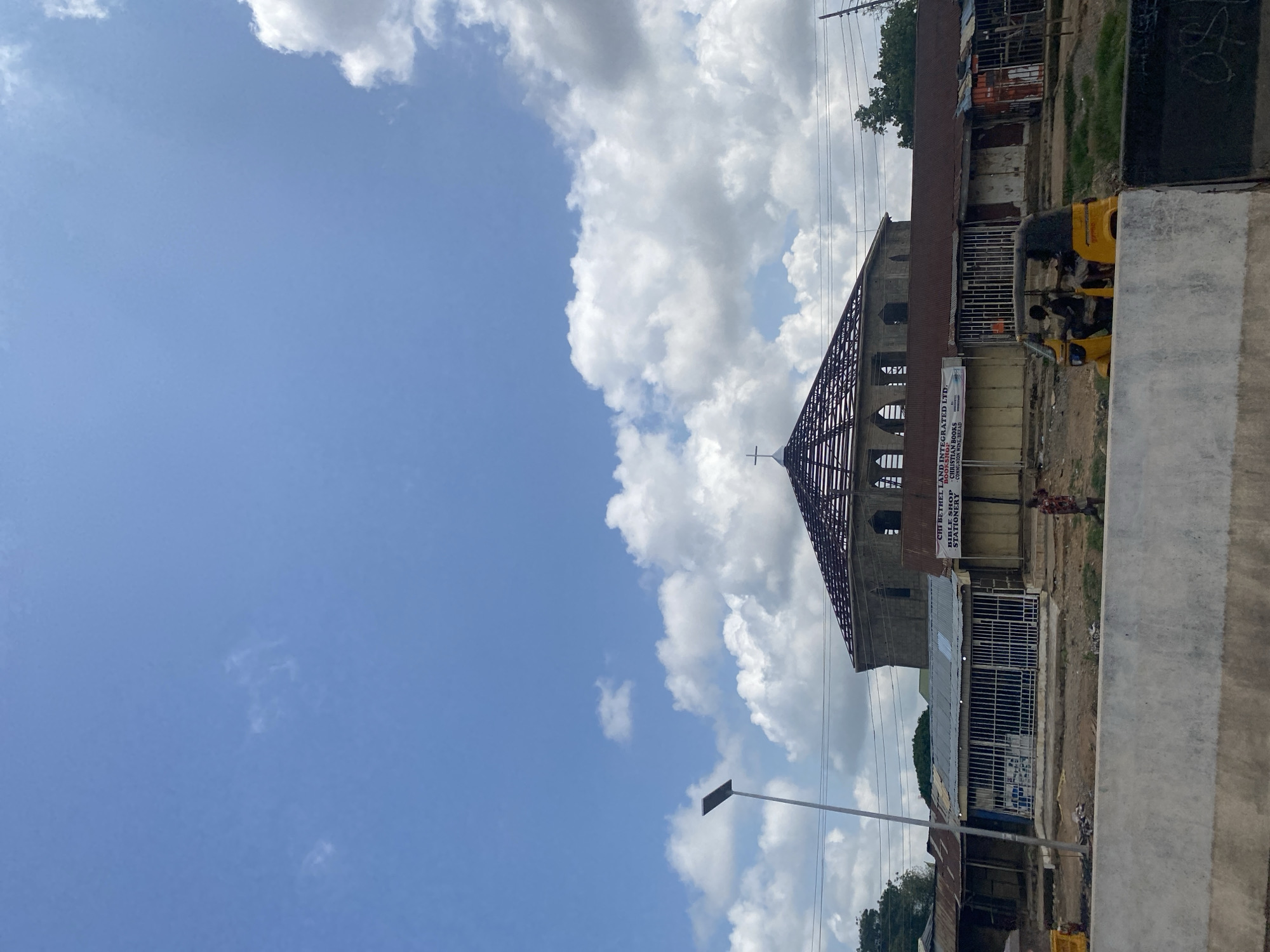 The image depicts a street scene in Ado, Nigeria, under a bright, partly cloudy sky. In the foreground, a concrete barrier runs horizontally across the lower part of the frame. Beyond the barrier, a dirt road is visible, lined with small shops and businesses with corrugated metal roofs. One shop has a sign that reads "CHI BETHEL LAND INTEGRATED LTD. BOOKSHOP STATIONERY." A yellow motorized tricycle, commonly known as a "Keke Napep" or "Marwa," with two passengers, travels on the road. Further back, a large, domed building with a cross on top, possibly a church under construction or renovation, dominates the background. The structure appears to be made of stone or concrete, with its roof framework exposed. Another street lamp is visible on the left side of the image. A single person is walking on the dirt road near the shops. The overall impression is a busy street scene in a Nigerian city, with a mix of commercial activity and ongoing construction.