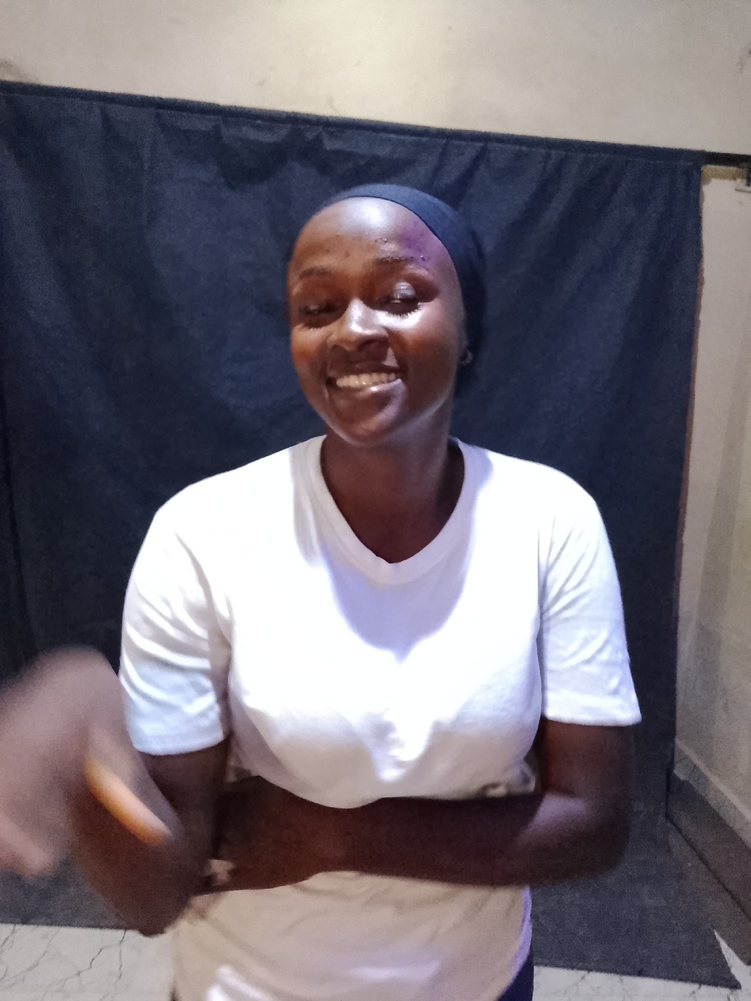 A woman with dark skin and a black headband smiles with her arms crossed in front of a dark blue curtain in an indoor setting. She wears a white t-shirt. An arm extends into the frame from the left side. The location is in the city of Unknown, Unknown.