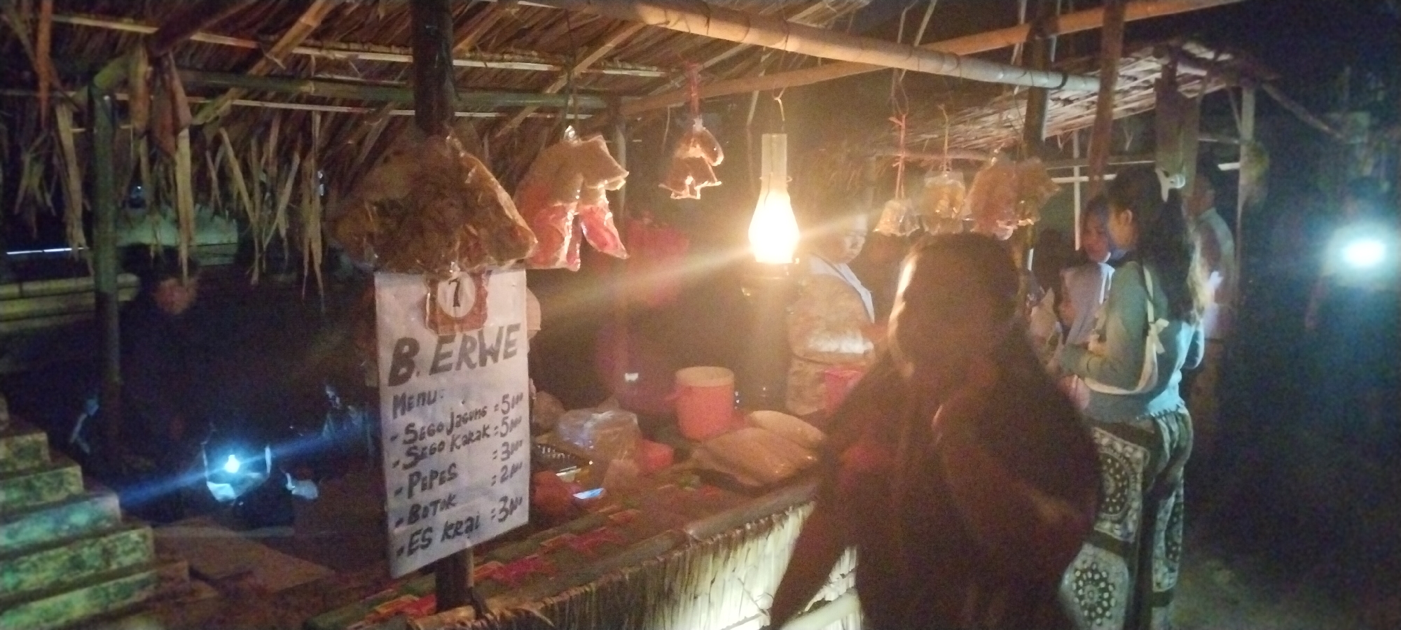 The image depicts a dimly lit outdoor market stall in Keting, Indonesia, at night. The stall has a thatched roof supported by bamboo poles, with dried vegetation hanging from the ceiling. Several items, possibly food items, are hanging from ropes. A sign displays "B. ERWE" and a menu with items such as "Sego Jagung," "Sego Kakak," "Pipes," "Botok," and "ES kreai" with prices. A kerosene lamp illuminates part of the stall. Several individuals are visible around the stall, some appearing to be customers and others possibly vendors. One person in the foreground is partially obscured. The overall scene suggests a local, informal market setting.