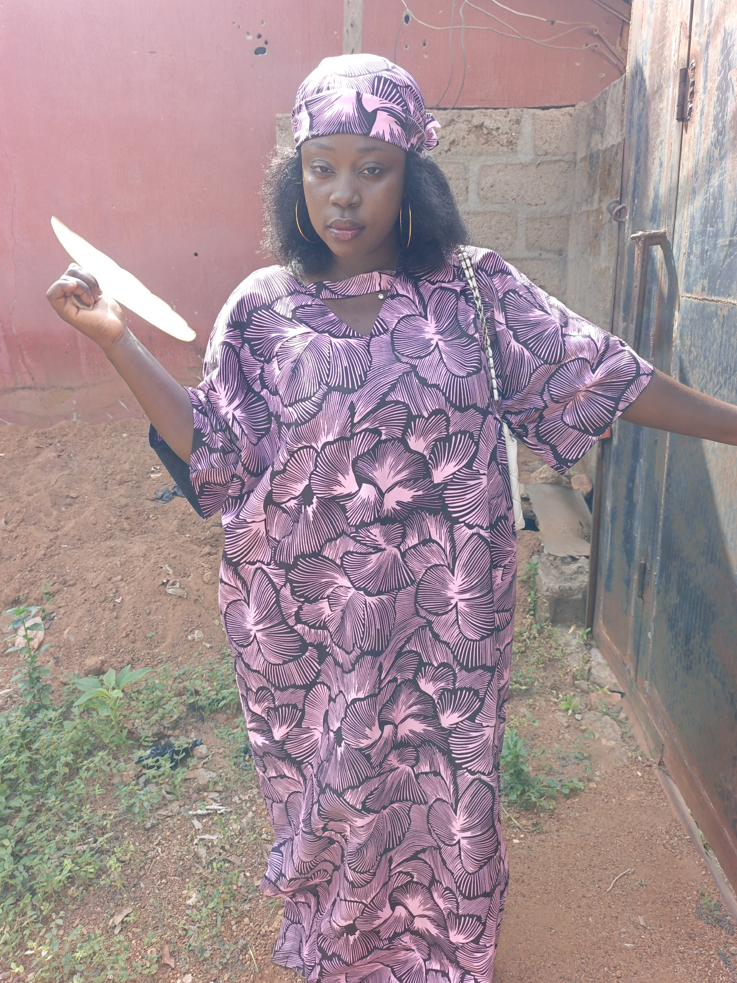 A medium shot captures a woman outdoors, possibly in Akwanga, Nigeria, given the context. She is wearing a patterned pink and black dress with matching headscarf and gold hoop earrings. She holds a light-colored, flat object in her left hand.
She stands on reddish-brown soil amongst sparse vegetation. Behind her is a red wall on the left and a section of a block wall and a weathered, gray metal gate on the right. An electrical wire is visible overhead.