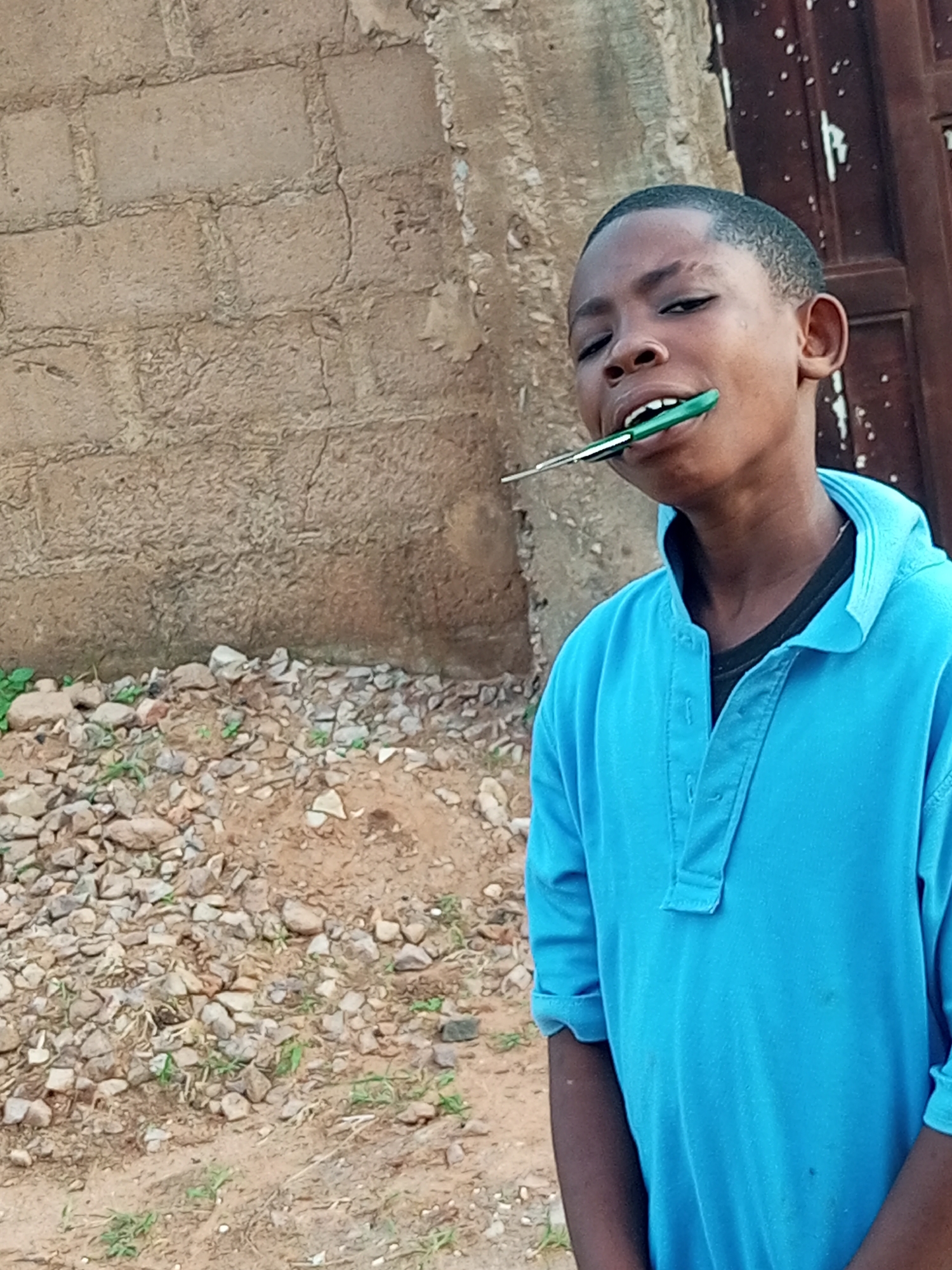 A young boy, appearing to be of African descent, stands outdoors in what could be the city of Tempure, Nigeria. He is wearing a bright blue collared shirt, unbuttoned at the top, revealing a black t-shirt underneath. His hair is short and black. The boy is holding a green and silver object, possibly a pen, in his mouth, with the green end angled upwards. His head is tilted back slightly and his mouth is open.
The background consists of a rough, tan-colored brick wall, and to the left, a pile of small stones and dirt with sparse green vegetation. To the right of the boy, a portion of a dark brown door or wooden structure is visible. The lighting suggests it is daytime.