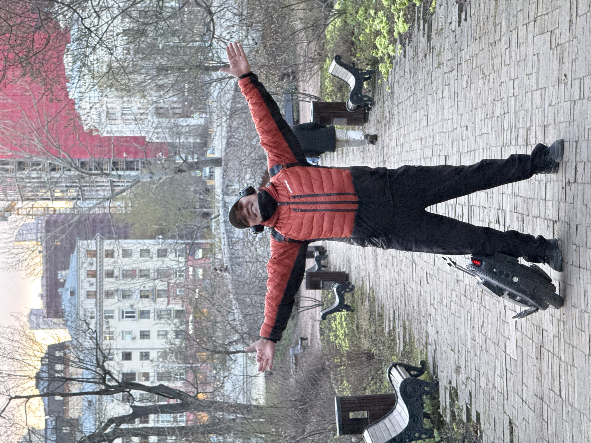 A man stands on a brick sidewalk, arms outstretched in a 'V' shape. He wears a red and black jacket, black pants, and a cap. A black object with red lights is near his feet. Several park benches line the sidewalk. In the background, buildings and trees are visible. The location is Moscow, Russia.