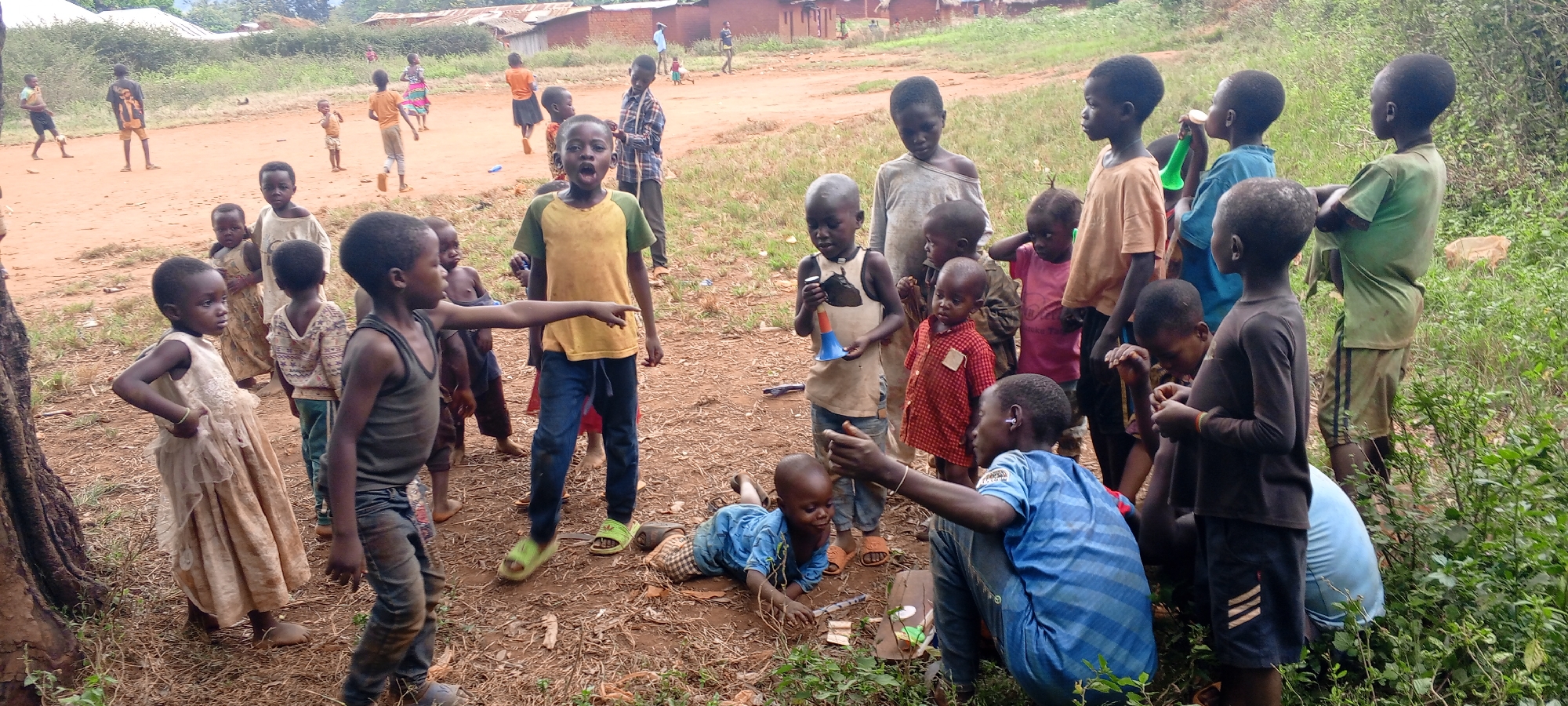 In a rural area of Ngalula, Democratic Republic of the Congo, a group of children, predominantly of African descent, are gathered in an open dirt area with sparse grass and vegetation. In the background are several small, simple structures that appear to be houses or buildings. In the foreground, a man is crouched, interacting with a toddler lying on the ground. Several children are standing and looking at the man and toddler. Some are holding small, colorful toys or instruments. Another child is pointing toward the background. The children are wearing a variety of casual, often worn, clothing. The overall scene suggests a spontaneous gathering or play activity in a village setting.