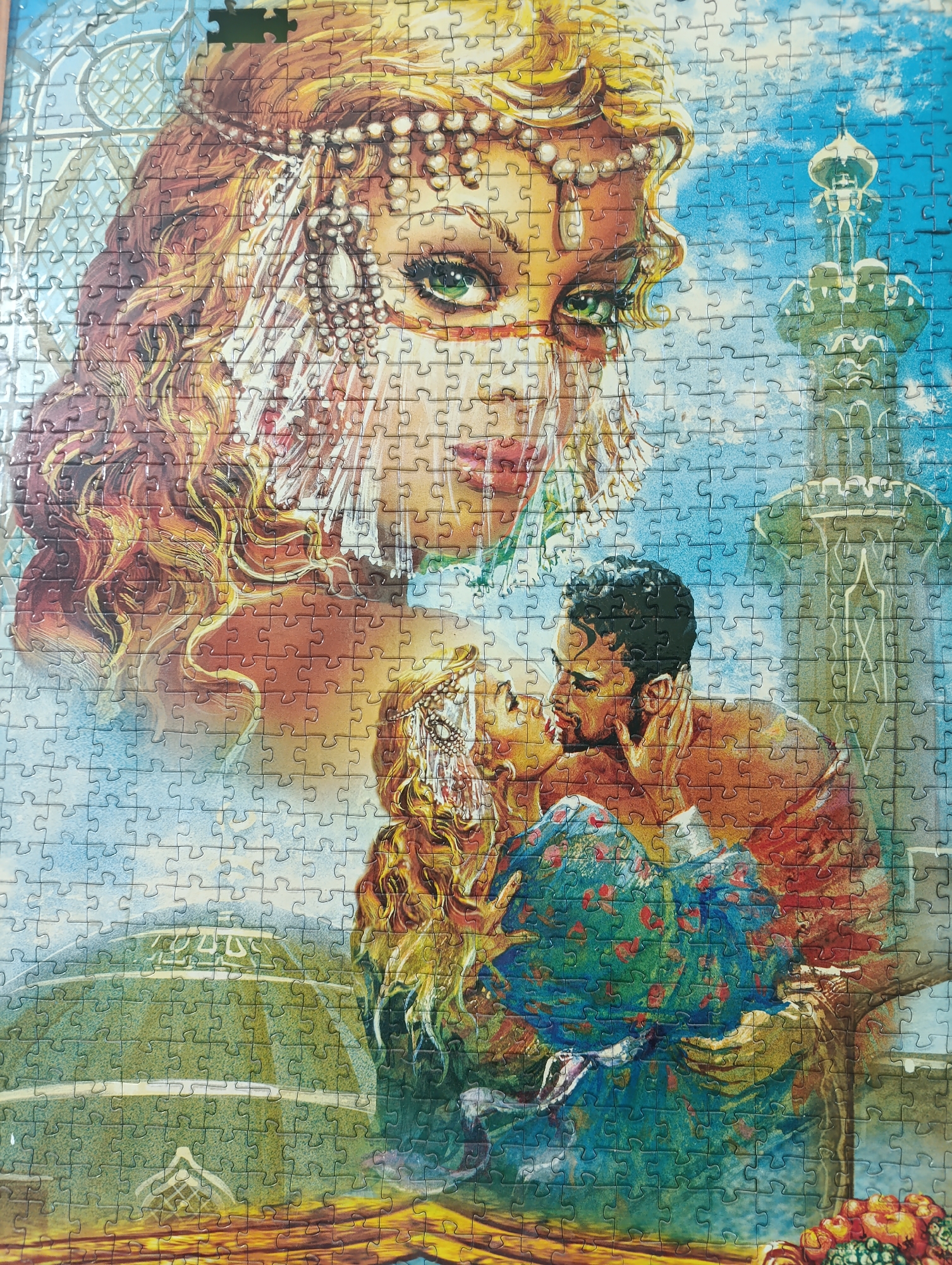 The image is a close-up, high-angle, overhead shot of a jigsaw puzzle depicting a scene with an ornate, large-scale portrait of a woman with flowing red hair and veiled jewelry, overlaying a romantic scene of a couple embracing. The woman in the portrait has striking green eyes and is adorned with a pearl and jeweled headdress and veil. Below her, a man and woman are kissing passionately, with the woman wearing a blue and green patterned garment and the man in an orange-red top. The background features elements that appear to be an architectural structure resembling a minaret or tower and a domed building, set against a vibrant blue sky with scattered clouds. The puzzle pieces are clearly visible, creating a textured, mosaic-like effect across the entire image. There is no indication of this scene being located in Moscow, Russia.