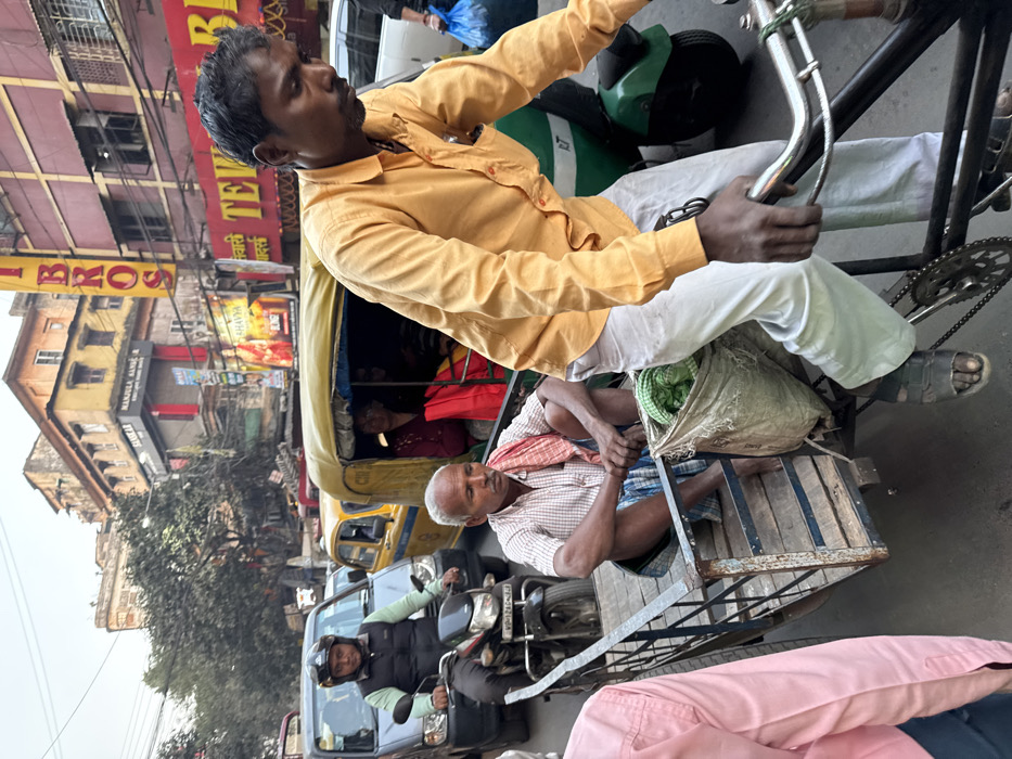 This image depicts a congested street scene in Kolkata, India, observed during daylight hours.
In the foreground on the right, a male bicycle rickshaw puller, dressed in a mustard-yellow long-sleeved shirt and light grey trousers, is actively pedaling his rickshaw. His hands grip the handlebars, and his right foot is visible in a dark sandal. An off-white sack, containing green fabric, hangs from the rickshaw's frame near his leg. Seated on the wooden platform of the rickshaw is an older male passenger, barefoot, wearing a light-colored checkered shirt and a blue and white patterned lungi. A red and white striped gamcha (towel) is draped over his left shoulder, and his hands are clasped between his knees as he looks forward.
In the mid-ground, a variety of vehicles are present in traffic. On the far left, a dark SUV-like vehicle is partially visible, followed by a man on a black motorcycle, wearing a black helmet and a light green shirt over a black vest. The motorcycle's license plate shows "MP-12." Behind the motorcycle, a yellow Ambassador taxi is visible, a common sight in Kolkata. To its right, a yellow auto-rickshaw contains two female passengers, one in a maroon top holding a red bag. A green electric auto-rickshaw is partially visible further back on the right.
The background features densely packed, multi-storied commercial buildings of varying age and architectural styles. Prominent signage includes a vertical yellow sign with red lettering spelling "T BROS," another sign reading "MANJULA SANGEET," and a board for "AHAVYA" displaying a saree figure and "10%." Below the "T BROS" sign, a red building features a partially visible sign starting with "TEWARI BE...". Numerous overhead electrical wires are visible crisscrossing the scene.
Based on the presence of yellow Ambassador taxis, traditional bicycle rickshaws, and distinct commercial signage like "Manjula Sangeet" (associated with Manjula Sangeet Karyalaya, a known musical instrument store), the location is identified as a busy commercial street in Kolkata, India, likely within an area such as Esplanade or Chandni Chowk.