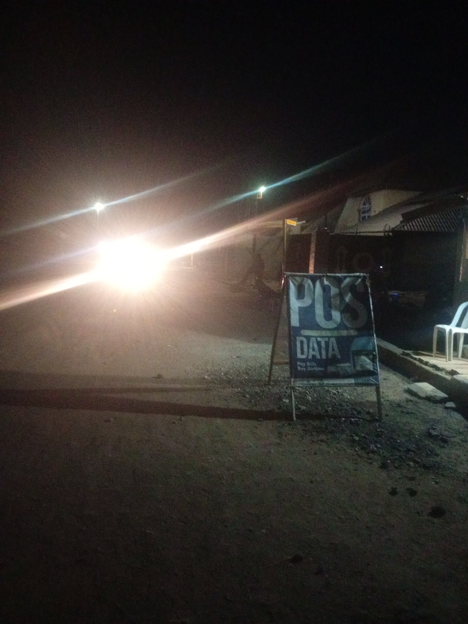 The image captures a nighttime scene on a street in Sabon Gari, Nassarawa, Nigeria. A bright, intense light source, likely from a vehicle's headlights, illuminates the left side of the frame, creating a strong glare. In the foreground, to the right of the main light source, an A-frame sign displays "POS DATA" prominently, with smaller text below indicating "Pay Bills Buy Airtime." The sign is dark blue with white lettering. Behind the sign, a dimly lit building with a corrugated metal roof is visible. To the far right of the image, a white plastic chair sits on a raised platform. The ground appears to be unpaved and uneven, with scattered debris. The overall atmosphere is dark, with the intense light and the sign being the primary points of focus. There are no people clearly visible in the image.