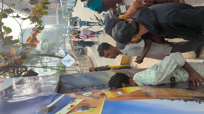 A group of people are on a street. Some people are lying down on the ground, while others are standing and looking at them. The people lying down appear to be posing or performing an activity. In the background, there is a street with vehicles and buildings. A tree with orange leaves is on the side.