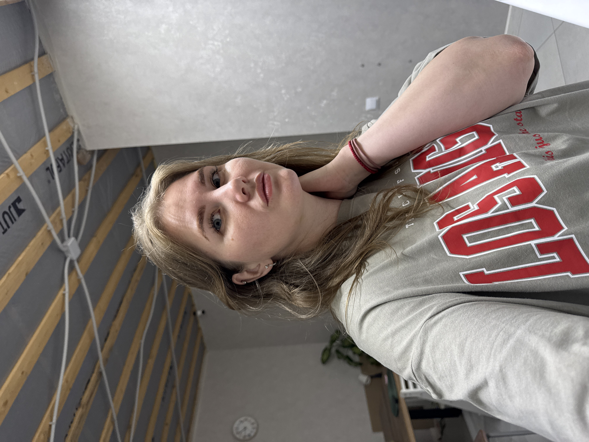 A woman is the primary subject of this portrait-style image. Her complexion is fair, with light brown hair and blue eyes. She wears a casual, long-sleeved gray shirt with a bold red inscription "LOS ANGELES" printed on it. A thin, multi-strand red bracelet encircles her left wrist. Her left arm is bent and positioned to support her neck.
The background is diverse, consisting of interior and structural elements. To the left, the wall appears to be in a state of construction or renovation, featuring gray panels accented by exposed wooden beams. White electrical wiring is also visible. The upper portion of the background is a solid light gray, suggesting a painted or finished wall.
Below the subject and the wall in progress is a more standard interior space. A circular white clock is on the wall. To the right, a potted plant is sitting on a cabinet.
The location may be in the city of Gornyi (Dvurechenskoe S/P), Russia. However, without more details, this cannot be confirmed.