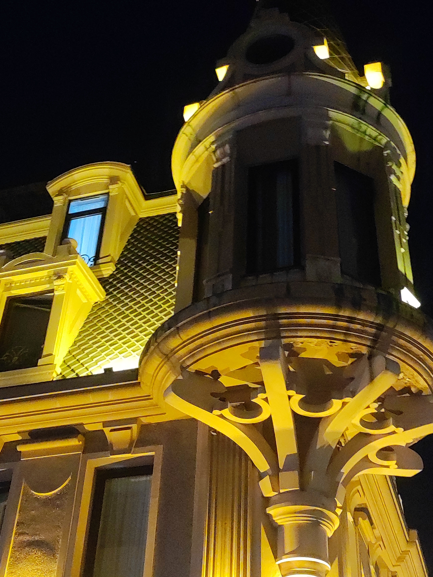 The image shows a close-up, low-angle view of the exterior of a building at night, illuminated by warm yellow lights. The architecture appears to be ornate, with a turret-like structure featuring decorative elements and several small lights along its upper edge. Below this, a curved balcony-like section is supported by decorative brackets. To the left, a portion of the building shows a tiled roof, a window with blue curtains, and another window with dark glass. The building's facade is detailed with molding and recessed panels. The overall impression is of an older, grand building, possibly a hotel or historic structure, located in Batumi, Georgia. There are no people or animals visible in the image, and the environment is dark, indicating nighttime.