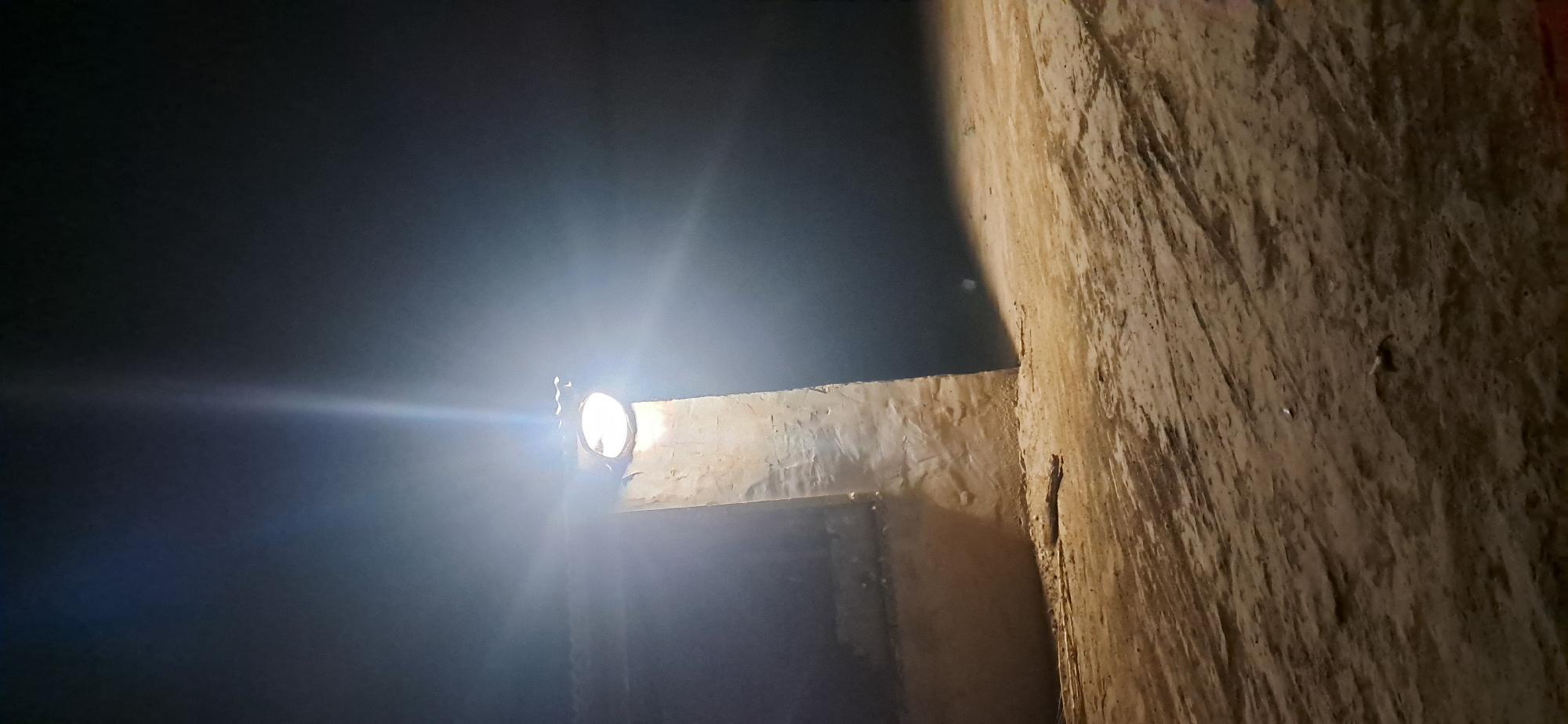 The image depicts a nighttime scene illuminated by a bright floodlight mounted on a rough, concrete-like pillar. The pillar is positioned in front of a darker structure with a textured wall. The ground in the foreground is sandy or dusty, with visible tire tracks or indentations. The floodlight casts a strong, direct beam upwards and outwards, creating bright highlights and casting shadows. The overall atmosphere is dimly lit, with the darkness of night surrounding the illuminated area. There is no visible presence of people or specific landmarks that would indicate the city of Yazy, Turkmenistan.