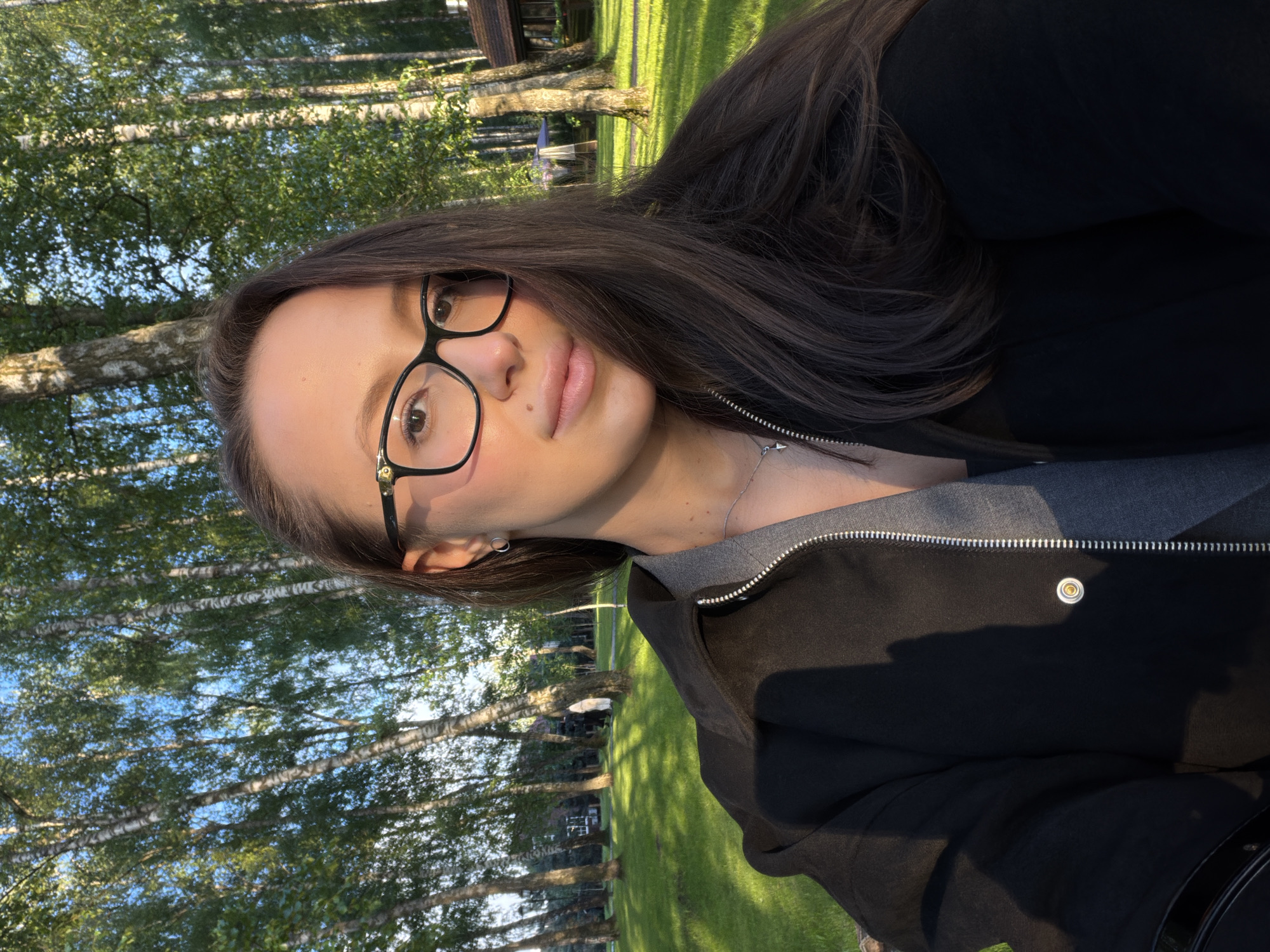 A woman with long brown hair and glasses is pictured outdoors in what appears to be a park or wooded area within Роса Снт, Russia. She is wearing a dark jacket over a gray top and has a necklace with a pendant. The background includes green grass, trees with white bark, and some glimpses of buildings in the distance. The overall impression is a natural setting with evidence of human habitation.