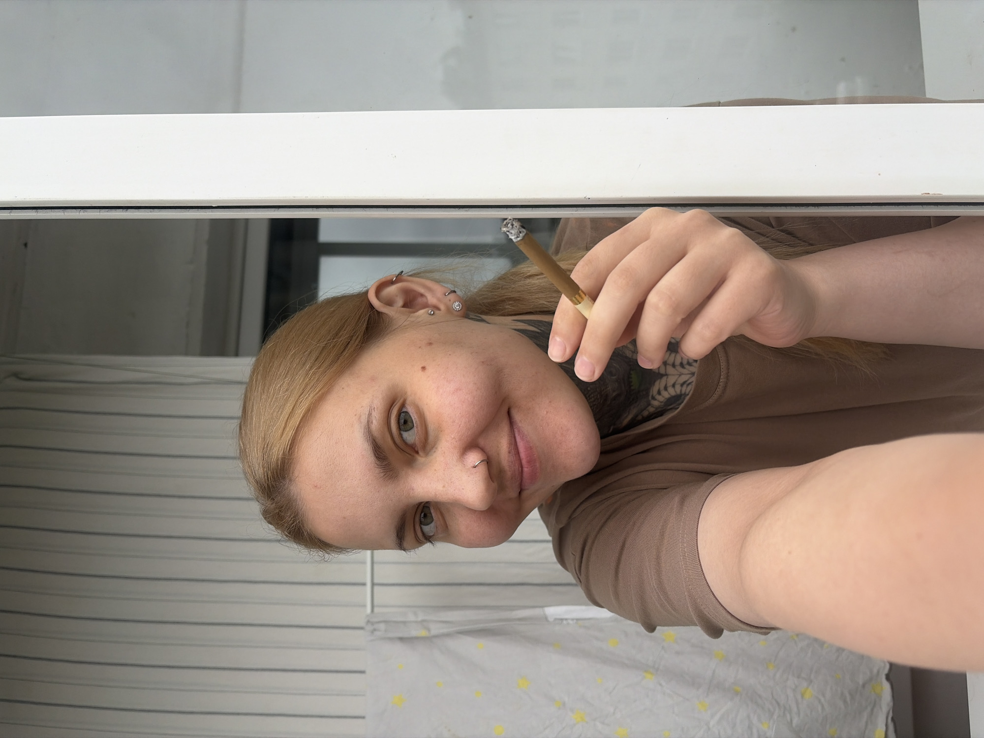 A blonde-haired person is holding a lit cigarette in their right hand, peering through a partially open white-framed window. The person has fair skin, a nose ring, and multiple earrings in their left ear. A tattoo is visible on their neck. They are wearing a brown t-shirt. The background shows white and grey striped curtains and a glimpse of an adjacent building. The location is Chaikovsky, Russia.