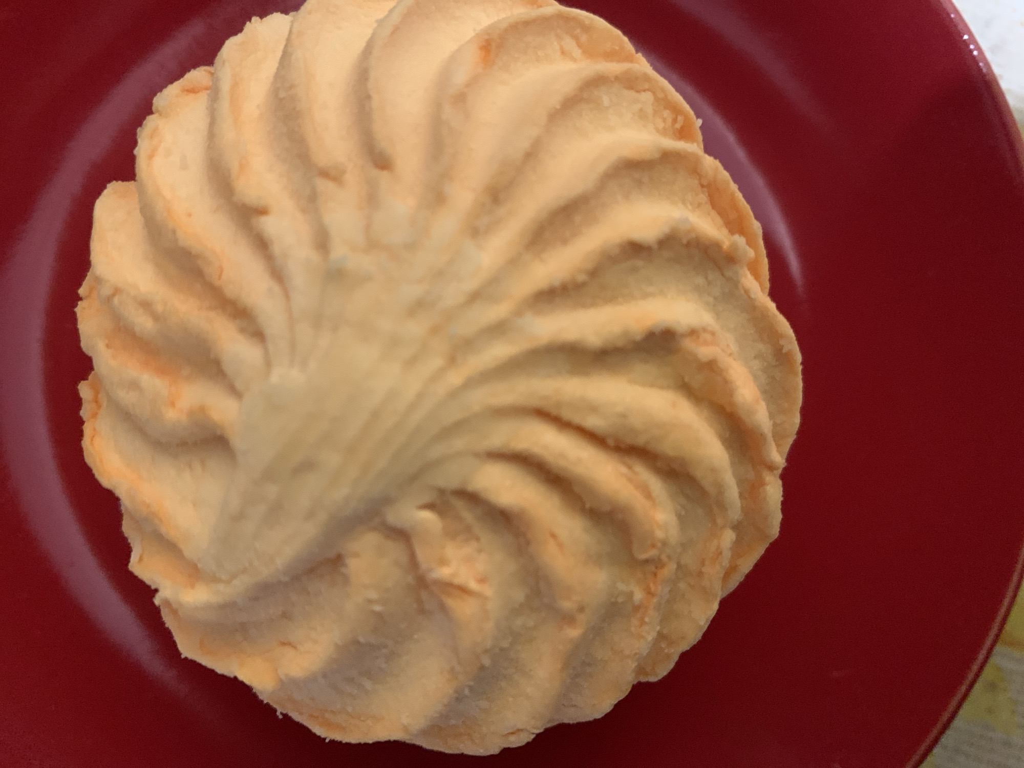 A single, round, orange-colored meringue sits atop a red plate. The meringue has a swirled, rippled texture radiating from the center. The plate rests on a surface with a pattern visible in the upper right corner. The location is Unknown, Unknown.