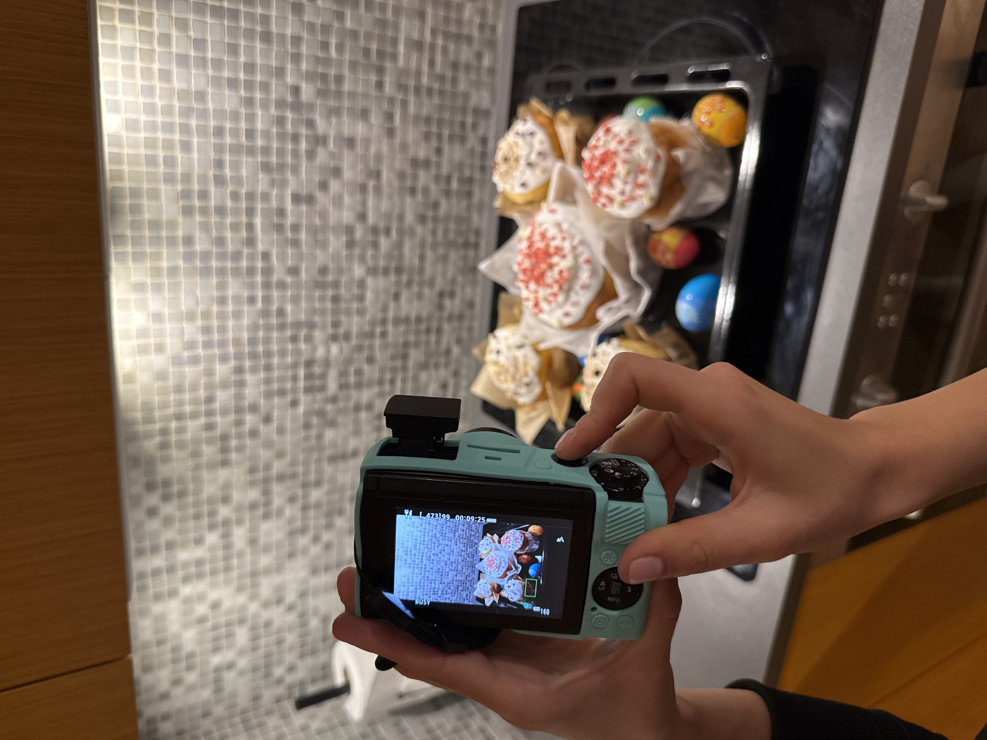 A person is holding a light blue camera, taking a picture of a tray of Easter cakes and colored eggs in what appears to be an oven. The camera's screen shows a similar image of the cakes and eggs, suggesting the person is reviewing a recent photo. The background features a mosaic tiled wall and a wooden-paneled wall. The context suggests this scene likely takes place within a kitchen, possibly in a home. There is no specific information to locate this in Warsaw, Poland.