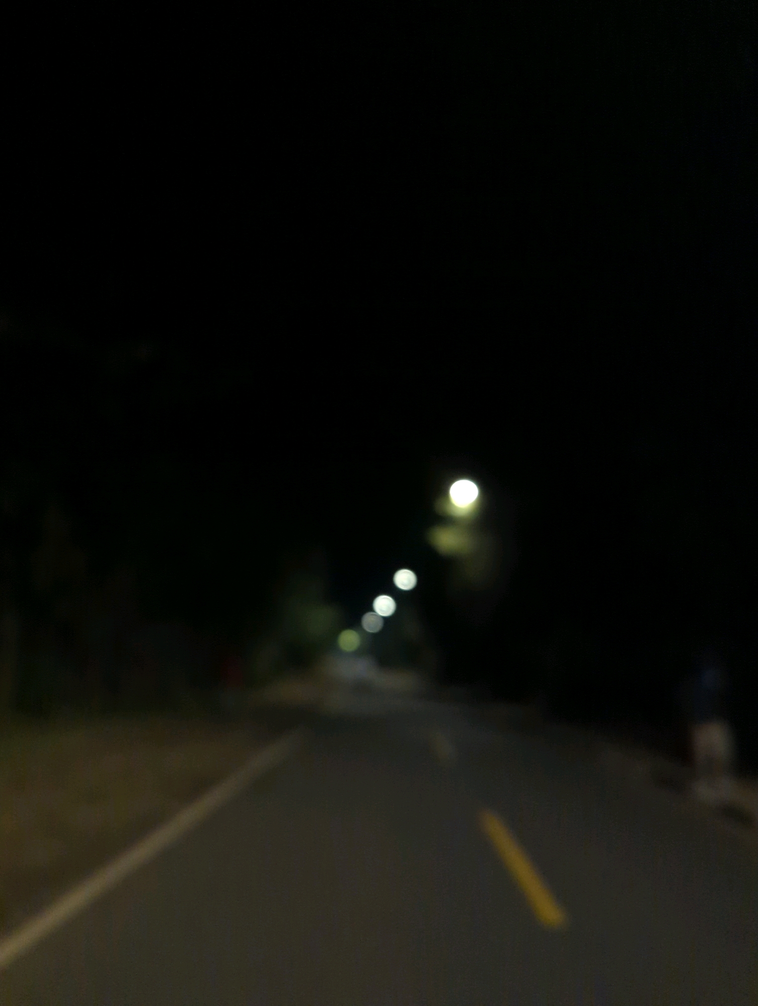 A blurred image captures a dimly lit asphalt road at night. Streetlights, appearing as out-of-focus circles of light, line the right side of the road, illuminating the dark surroundings. A yellow painted line runs down the center of the road. The left side of the road features a gravel shoulder, with the dark silhouettes of trees visible in the background. The overall impression is one of a quiet, possibly rural or suburban, street in Musanze District, Rwanda, at night.