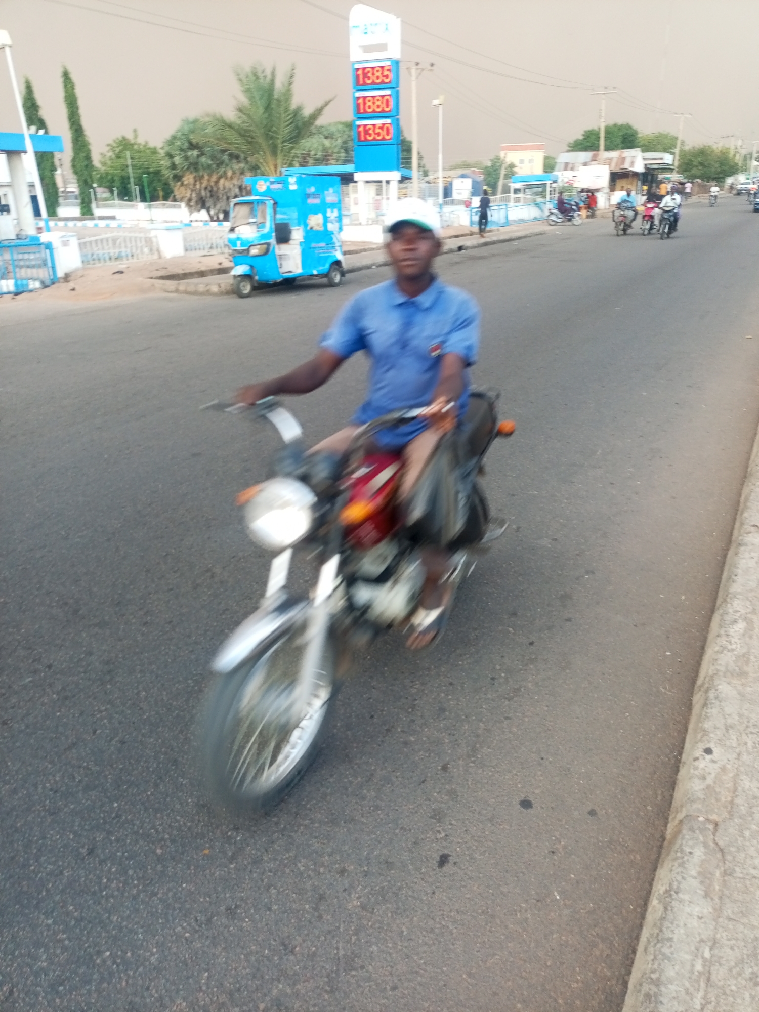 A man on a red and silver motorcycle, wearing a blue shirt and white cap, is riding on an asphalt road. Behind him are other motorcycles. A blue "Keke Napep" three-wheeled vehicle is parked near a Matrix gas station sign displaying prices: 1385, 1380, and 1350. The location is Gombe, Nigeria.