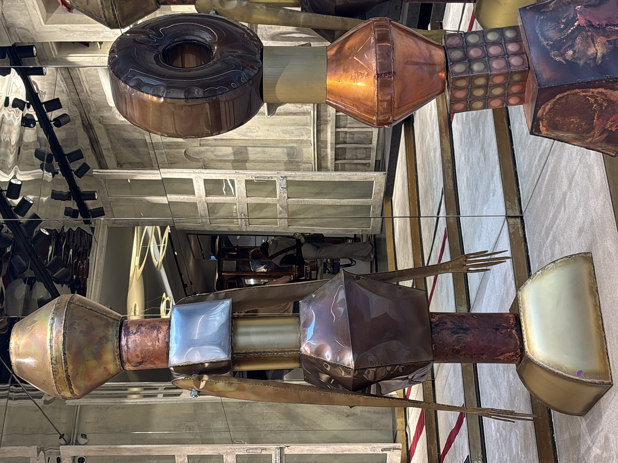 The image captures a detailed view of a multi-component sculptural art piece, suspended horizontally and largely reflected in a mirrored ceiling. The artwork is composed of various metallic and possibly plastic elements, joined linearly to form an elongated, complex structure.
The sculpture features distinct sections: a large, dark, glossy donut-shaped object, possibly inflatable, connects to cylindrical and conical sections in brass and copper tones. A cubic segment with a grid of smaller square or circular inlays in varied, muted colors (pink, yellow, green, brown) follows, leading to a darker, oxidized copper or bronze-toned cube. Further along, another conical section, transitioning from brass to copper, is attached to a silver-toned, reflective cubic part. From a dark, cylindrical segment, two skeletal, multi-pronged metallic "arms" extend downwards. The sculpture culminates in a pale, golden-toned, scoop-shaped or shield-like form.
The mirrored ceiling above reflects the underside of the sculpture and parts of the surrounding interior space. These reflections reveal a dark metal truss structure fitted with multiple spotlights, an industrial-style concrete ceiling, and rectangular window-like features. In the background, a person wearing light pants and a dark top is visible in the reflection, suggesting an indoor public or exhibition space. The setting is likely an art installation or a decorative element within a commercial or public building in Milan, Italy. There is no visible text.
