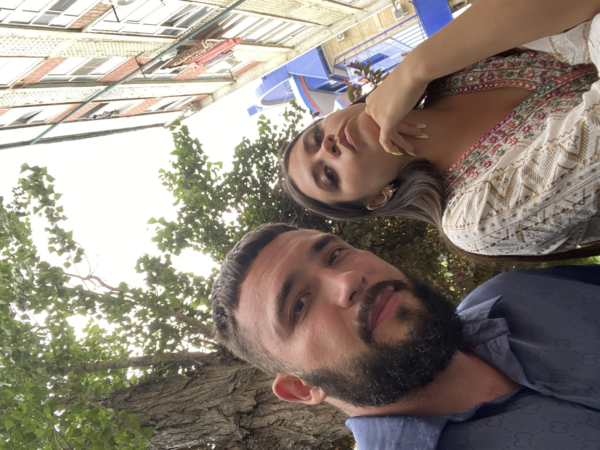 This is a selfie taken outdoors in the city of Derbent, Russia. The image features two individuals, a man and a woman, positioned in the foreground. The man, on the left, has short dark hair, a full beard, and is wearing a dark blue collared shirt with a patterned texture. He is looking directly at the camera with a neutral expression. The woman, on the right, has long dark hair and is wearing a white top with intricate colorful embroidery around the neckline and sleeves. She is resting her chin on her hand, also looking towards the camera with a contemplative expression.
The background reveals an urban environment. To the left, a large tree with textured bark and green foliage dominates the upper portion of the frame. To the right, a multi-story building with cream-colored brickwork, red accents, and numerous windows is visible. A section of a blue structure, possibly part of the same building or an adjacent one, is also present. The sky above is overcast, with bright white light suggesting daytime. The perspective of the selfie is looking upwards, contributing to the dynamic composition.