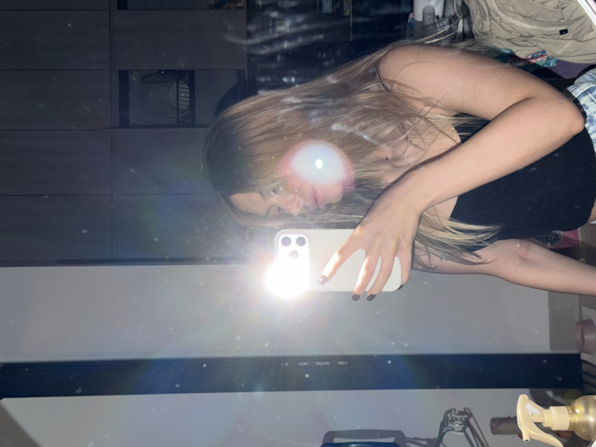 A young woman with blonde hair is taking a selfie in a mirror. She is wearing a black tank top and denim shorts. Her phone is in her right hand, and she is holding it up to take the picture. The flash on her phone is on, creating a bright light that illuminates the scene. The background appears to be a room with cabinets and shelves. The image has a reflective quality, possibly due to the mirror or the lighting. The location is not specified in the image.